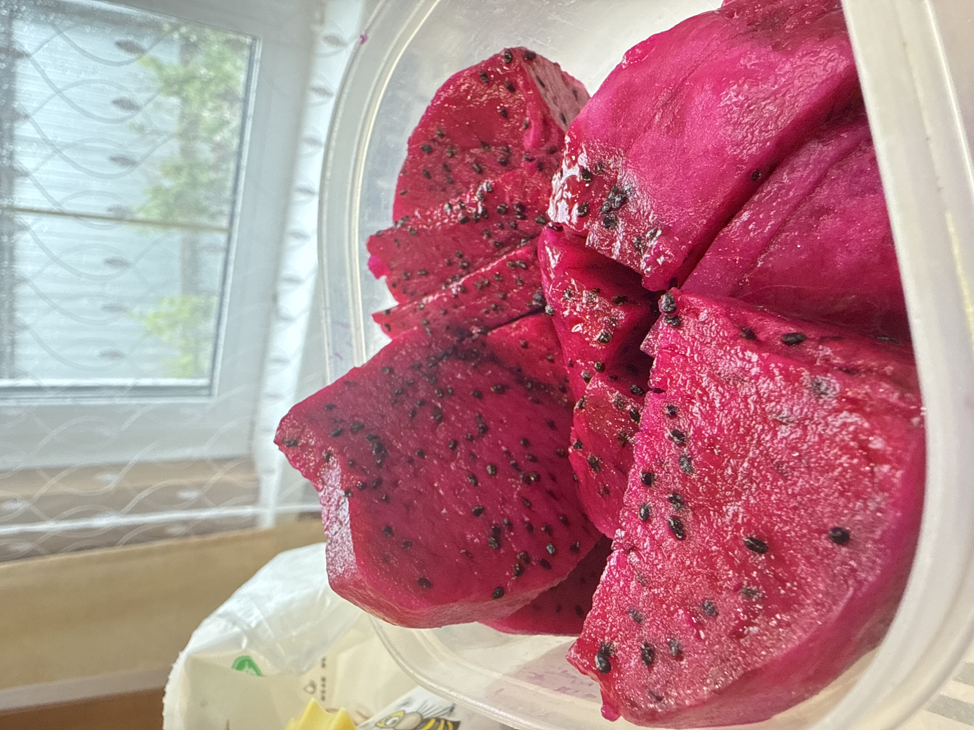 The image captures a close-up, slightly elevated view of vibrant, sliced red dragon fruit (pitaya) contained within a clear plastic container. The fruit's flesh is a deep magenta color, speckled with numerous small, black seeds. Several pieces of the fruit are visible, cut into thick, irregular slices, and appear moist.
In the background, a window provides natural light and a blurred view of the outdoors. Through the window, green foliage from trees or bushes is discernible, suggesting an outdoor environment. The window itself has a textured or patterned glass, possibly frosted or with an etched design, creating a somewhat diffused view.
Below the container and extending into the lower left corner, a crumpled white plastic bag or packaging is partially visible. On this bag, faint, blurry dark text and a small green symbol, possibly a 'V' shape, can be seen. To the immediate left of the bag, a sliver of a yellow object, possibly another piece of fruit like pineapple or a snack, is present, with indistinct cartoon-like facial features (eyes, mouth) and text barely visible on it.
The scene appears to be indoors, likely in a kitchen or dining area, during daylight hours, given the natural illumination from the window. The primary activity depicted is the preparation and storage of the fruit. No people are visible, and no specific emotions are conveyed.