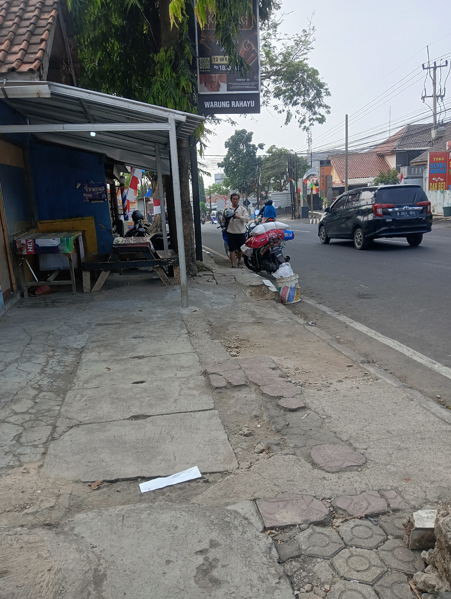 A street scene in Ciporang, Indonesia, shows a sidewalk beside a road. A man stands near a parked motorcycle, holding a helmet. To the left, a small, covered structure with tables and chairs appears to be a food stall. A black car drives on the road in the background. The sky is overcast.