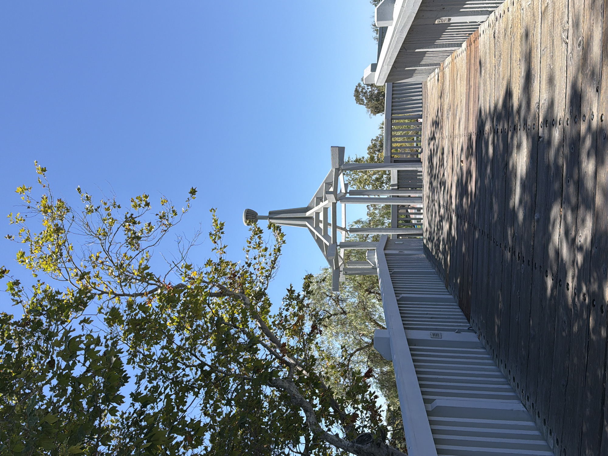 The image depicts a wooden pedestrian bridge with light grey railings, leading towards a light grey gazebo structure. The bridge is made of weathered wooden planks with visible nail heads. Sunlight casts shadows across the bridge from the surrounding trees. On the left side of the bridge, a large tree with green and yellowing leaves extends into the frame, reaching towards a clear blue sky. The background reveals more trees and a hint of distant foliage. The scene appears to be in an outdoor public space, likely a park or nature trail. No people are visible in the image.