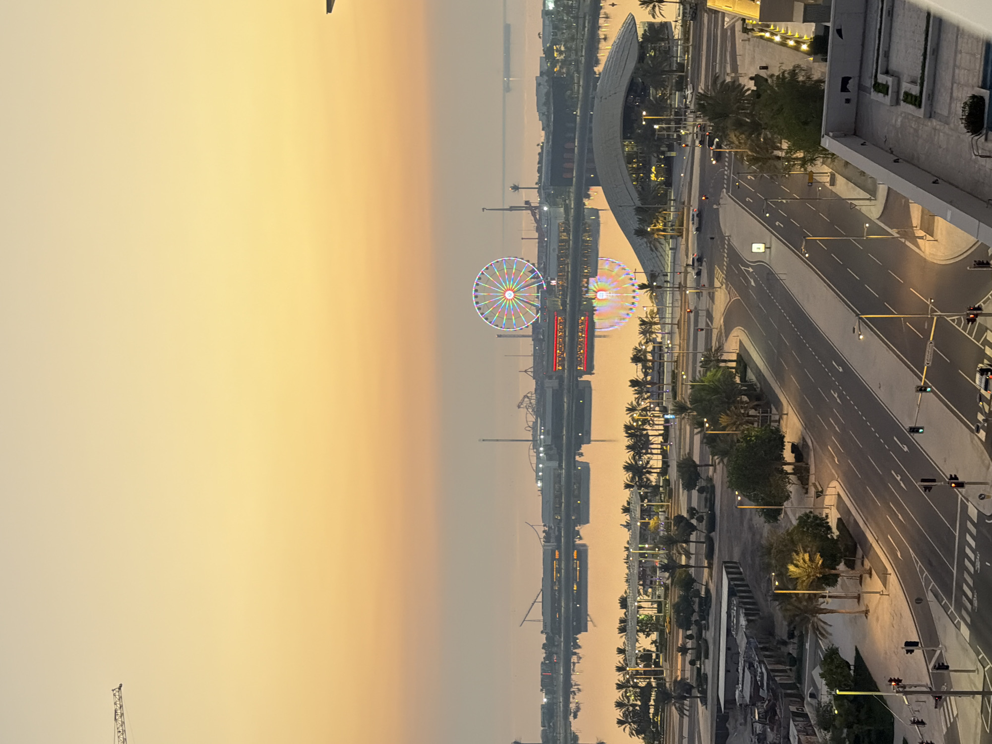 A long shot of a waterfront amusement park at dusk, with a Ferris wheel illuminated with colorful lights. The Ferris wheel reflects in the calm water, and a modern building with a curved roof is visible in the foreground. Palm trees line the waterfront, and a road with streetlights and traffic lights is in the lower portion of the image. The sky is a gradient of orange and yellow, indicating sunset. A construction crane is visible in the upper left corner of the image.