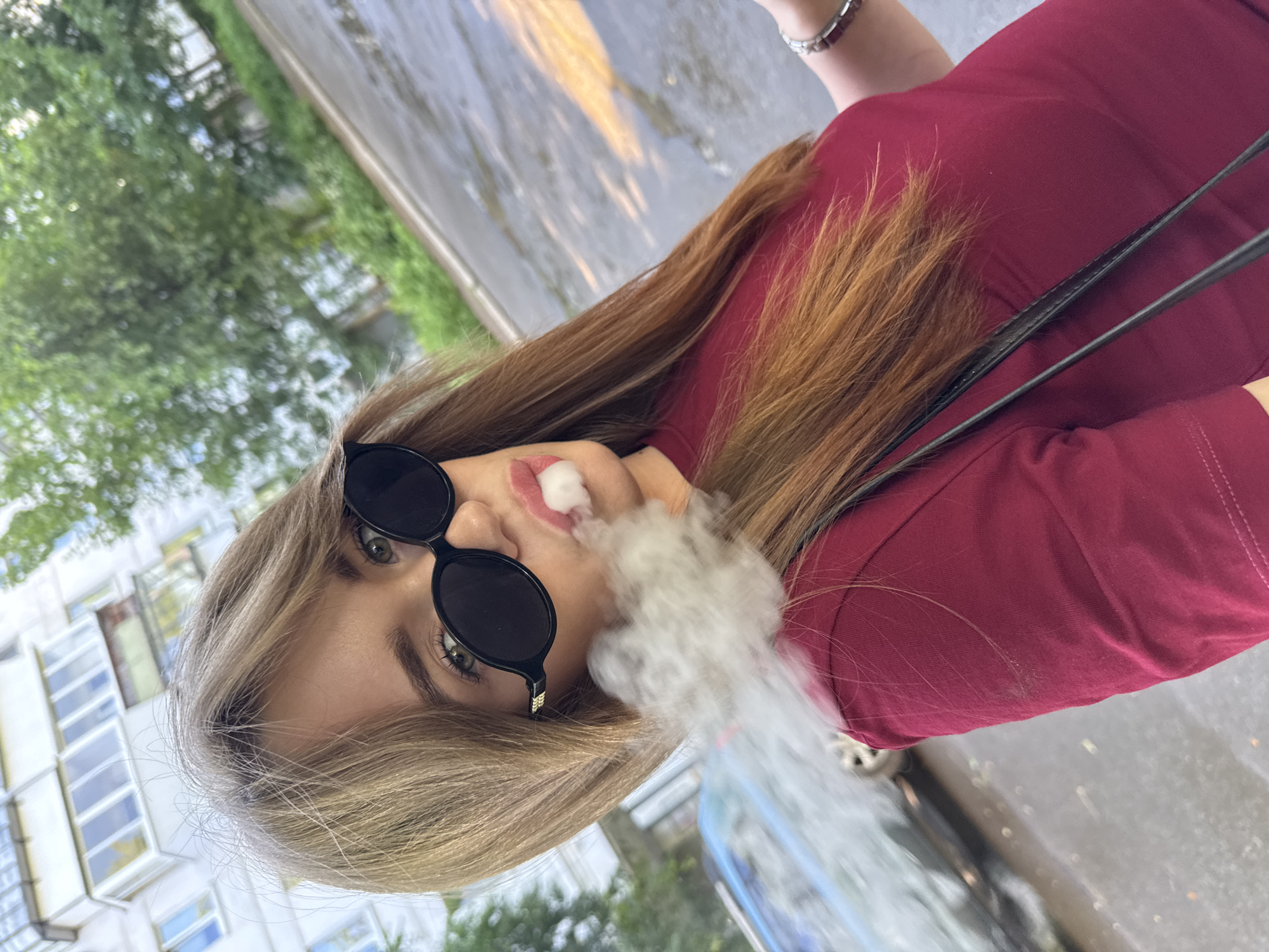 A young woman with long, reddish-brown hair and green eyes is shown in a close-up, selfie-style shot. She is wearing large, black, oval-shaped sunglasses and a maroon-colored top. She is exhaling a cloud of white vapor, likely from an electronic cigarette, which obscures part of her face and the foreground. The background appears to be an urban environment, possibly a street or sidewalk, with a wet pavement suggesting recent rain. A portion of a light-colored apartment building with multiple windows is visible in the upper left quadrant, and trees line the background. A blurred car is also visible on the left side of the image. The overall lighting suggests an outdoor setting during the daytime. There is no specific visual information in the image that places the scene in the city of Chaikovsky, Russia.