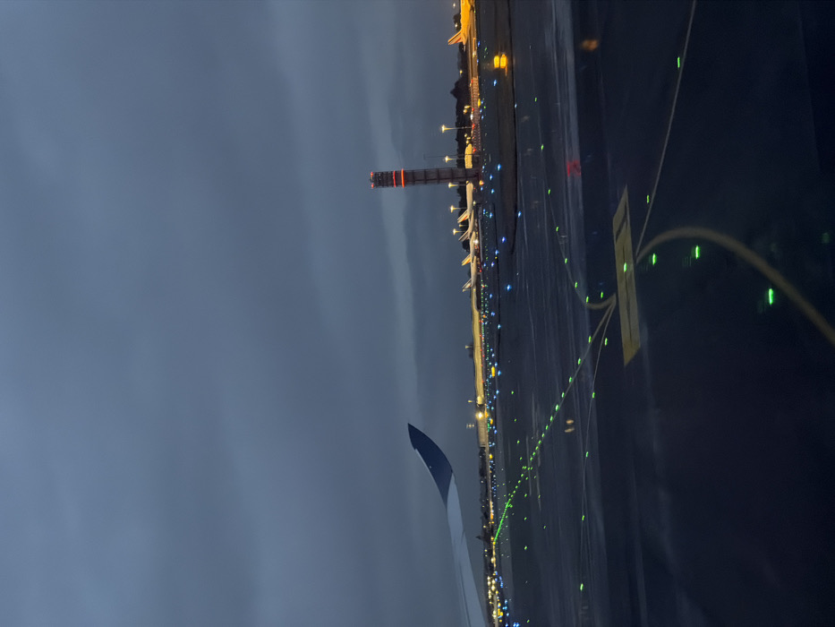 The image depicts an airport scene at night or late dusk, viewed from the window of an airplane on the ground. The sky in the upper left is dark and overcast. The primary subject is the illuminated airport tarmac or taxiway, which extends from the foreground towards the right side of the image.
The ground appears wet, reflecting the numerous artificial lights that define the airport infrastructure. Green lights are prominently displayed, outlining taxiway edges and guiding paths, with some forming curved lines in the mid-ground. Scattered blue lights are also visible, likely marking other sections of the airport. In the distance, yellow and orange lights illuminate various airport buildings and what appear to be parked aircraft.
A tall, distinctive tower with red and white horizontal stripes stands in the middle distance on the left side of the frame, emitting its own red warning lights. This is likely an air traffic control tower or a similar navigational structure. A portion of the airplane's wingtip, dark blue-grey, is visible in the lower left corner, establishing the passenger's perspective.
On the ground in the mid-right, a yellow rectangular marking with dark, indistinct text or symbols is visible, typical of airport signage. The overall atmosphere is that of a bustling yet quiet airport during non-daylight hours, with the wet surfaces adding a reflective quality to the scene. The location context for this active airport environment is Mitry-Mory, France.