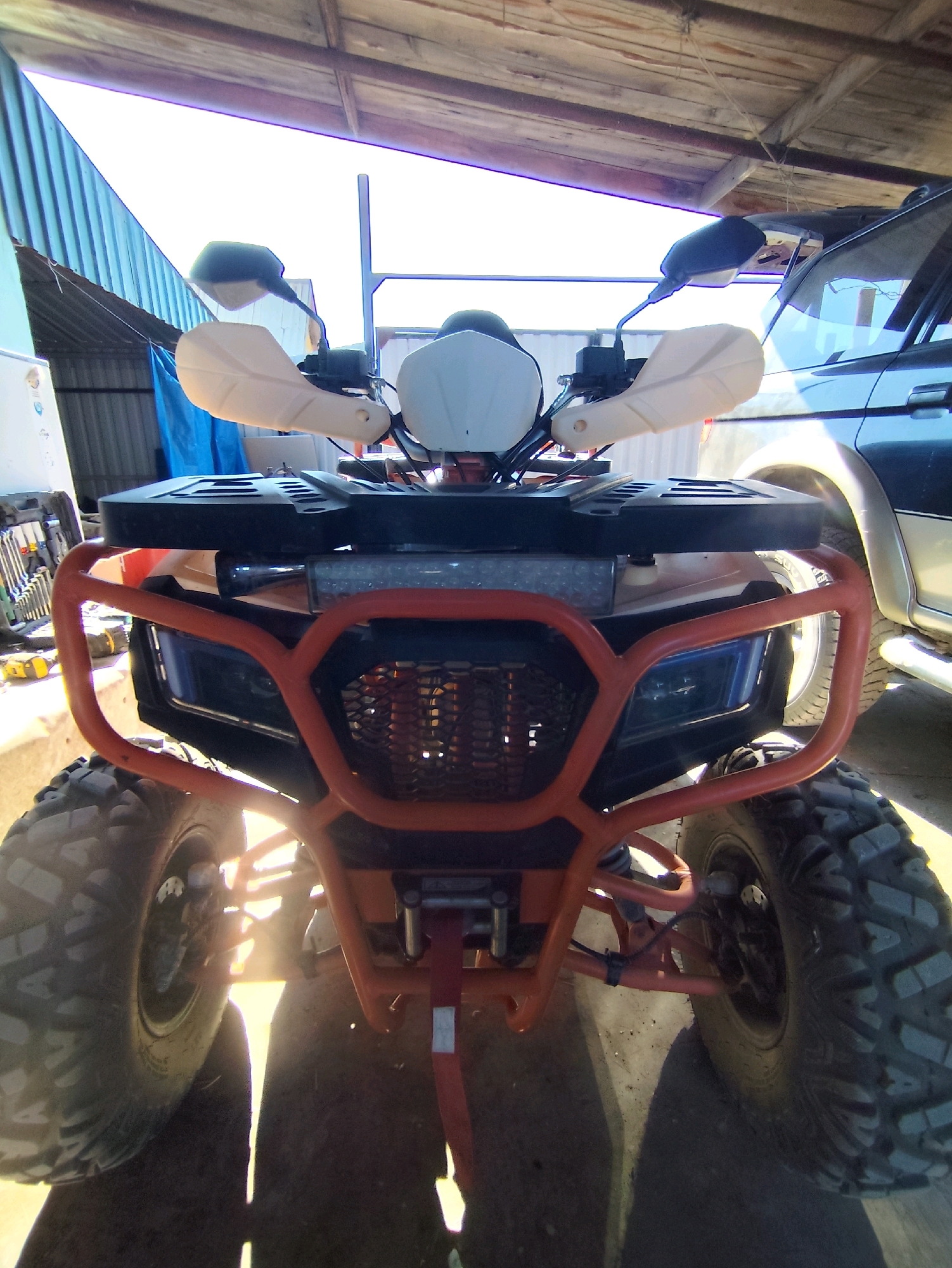 The image shows the front view of an orange and black ATV parked under a wooden and corrugated metal roof structure. The ATV has large, knobby tires, a prominent orange front bumper, and black handlebars with white handguards and black mirrors. A black rack is mounted above the front bumper. In the background, a dark-colored SUV is partially visible, parked to the right of the ATV. The environment appears to be a workshop or storage area, with tools and a blue tarp visible on the left. The lighting suggests it is daytime, with sunlight casting shadows. The location is specified as Balqaş, Kazakhstan.