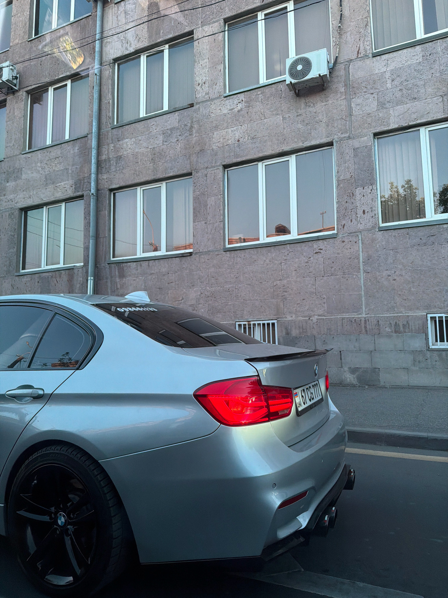 The image shows the rear passenger side of a silver BMW sedan parked on a street in Yerevan, Armenia. The car has tinted windows and black rims. The building behind the car is made of textured stone and has multiple windows. An air conditioning unit is mounted on the building's facade. A license plate reading "67 CG 777" is visible on the car's rear. The street appears to be paved, and there are some street markings visible. The lighting suggests it is daytime.