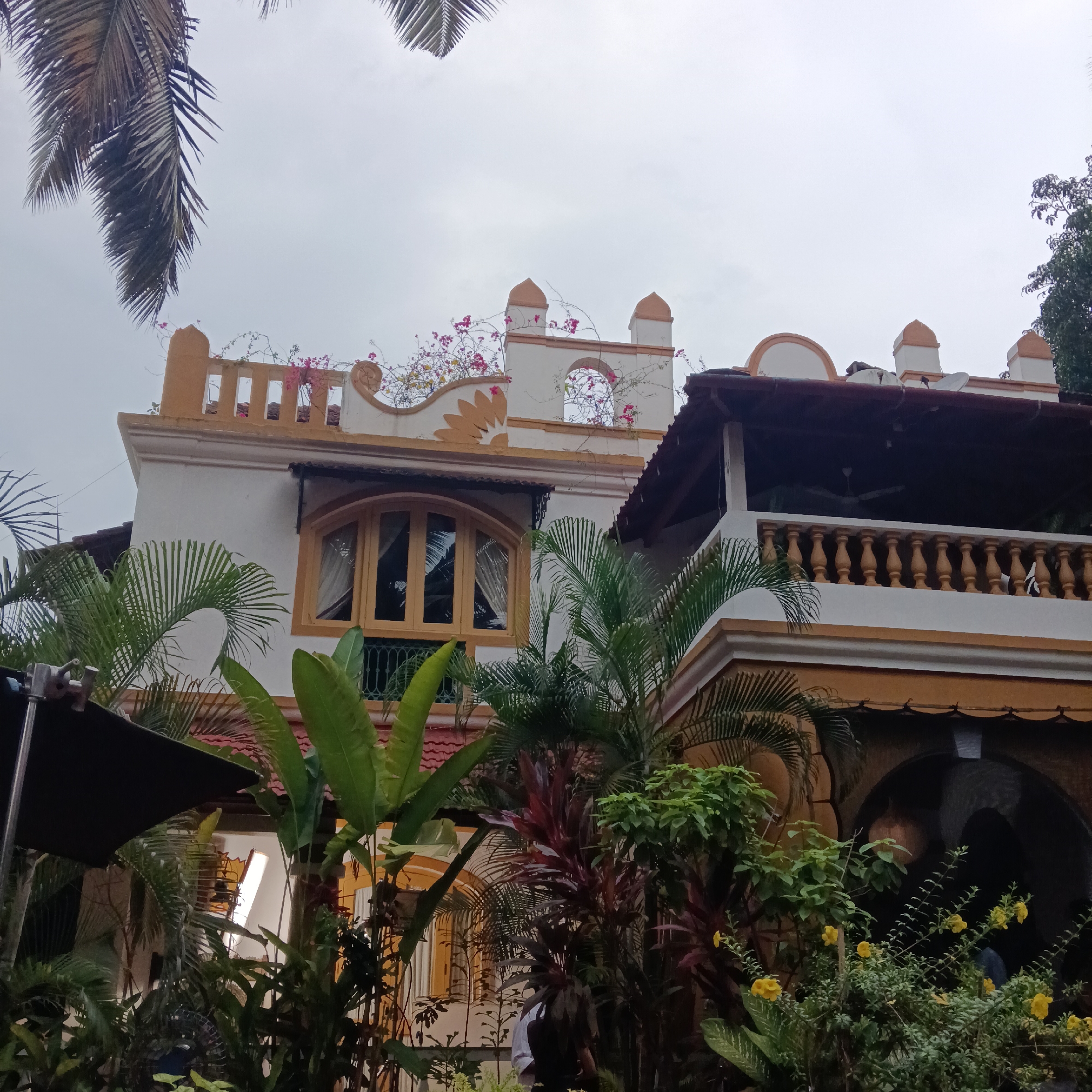 The image captures a building in Anjuna, India, characterized by its white facade with ochre trim and architectural details. The structure has a two-story section with a curved arch window and a balcony featuring a balustrade. Plants and trees, including palms and flowering bushes, densely populate the foreground, partially obscuring the lower portion of the building. A segment of a light-blocking screen is visible on the left, possibly indicating a film or photo shoot in progress. A couple of individuals are present in the lower portion of the building, with some portion of their body and head visible between the plants. The sky is overcast.