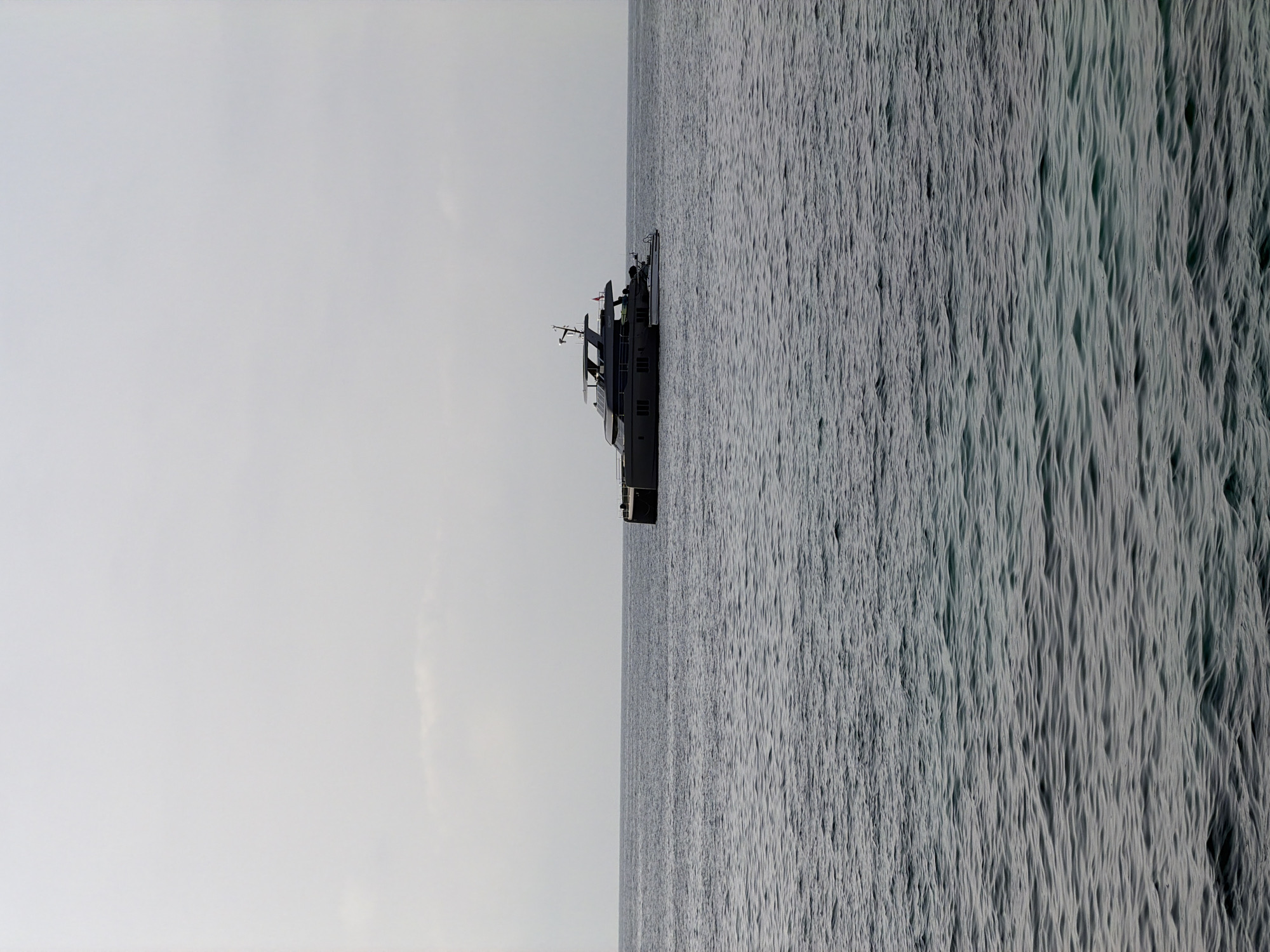 A dark-hulled luxury yacht is anchored in the calm, choppy waters of the Mediterranean Sea off the coast of Calvià, Spain. The sky above is overcast with a pale, hazy light. The yacht, featuring multiple levels and a sleek design, is the sole subject on the vast expanse of water, its reflection barely visible on the surface. The foreground consists of gently undulating waves. The overall scene conveys a sense of tranquility and isolation.