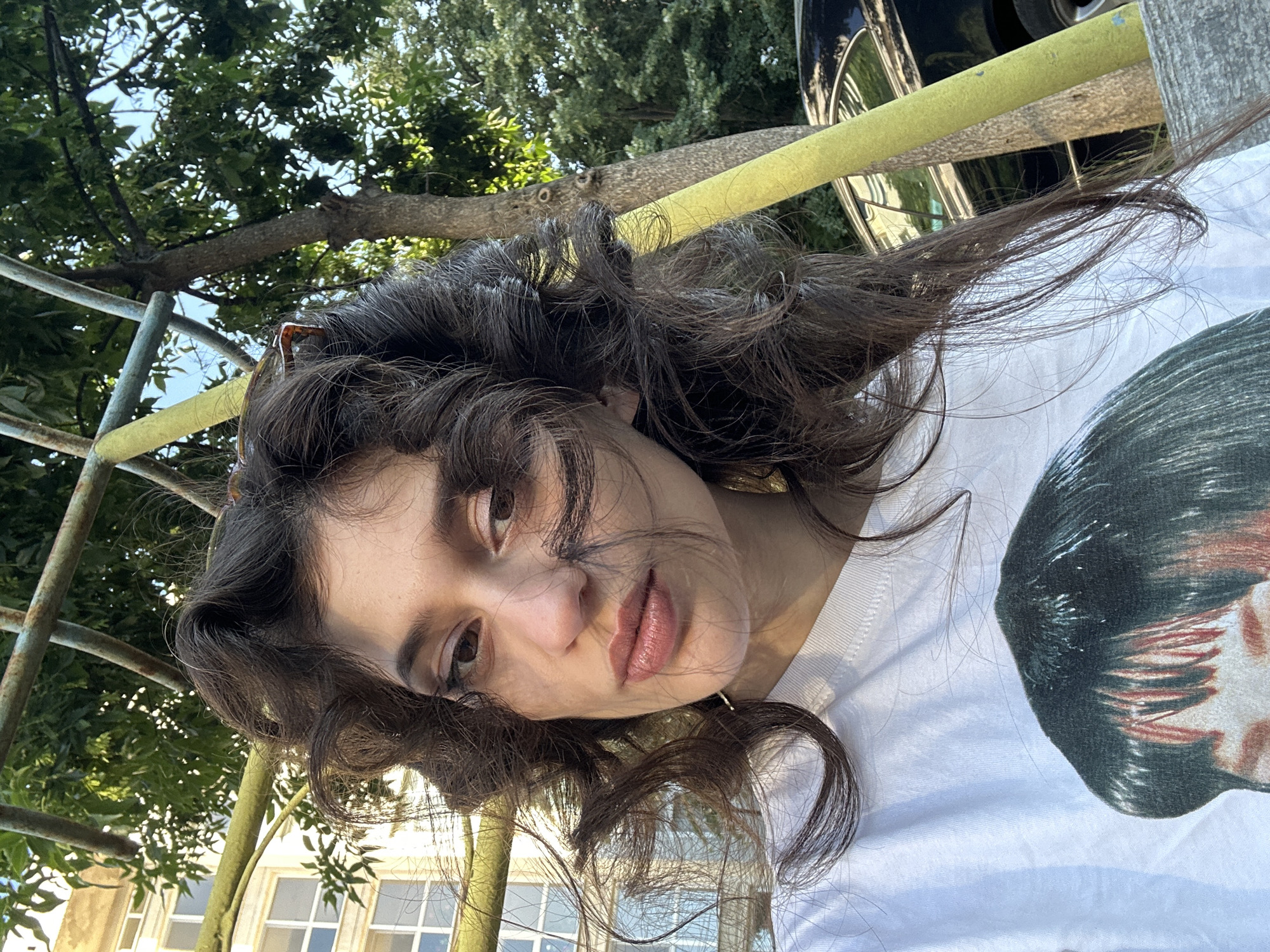 A young woman with dark, curly hair is lying down outdoors, with the camera angled from above. She is wearing a white t-shirt with a graphic print of a person's face on it. Her hair is messy and partially covers her face, with some strands blowing in the wind. She has light brown eyes and appears to be wearing light makeup. Her lips are slightly parted and appear to be glossed. Her gaze is directed towards the camera.
The background shows a tree with green leaves and a brown branch. A yellow railing runs horizontally behind the woman, and a metal structure, possibly part of a playground or outdoor seating, is visible to the left. Further in the background, there are glimpses of a building with windows and trees, suggesting an urban or park-like setting. The lighting suggests it is daytime, possibly late afternoon due to the warm tones. The overall mood is casual and relaxed. There is no visible text in the image.