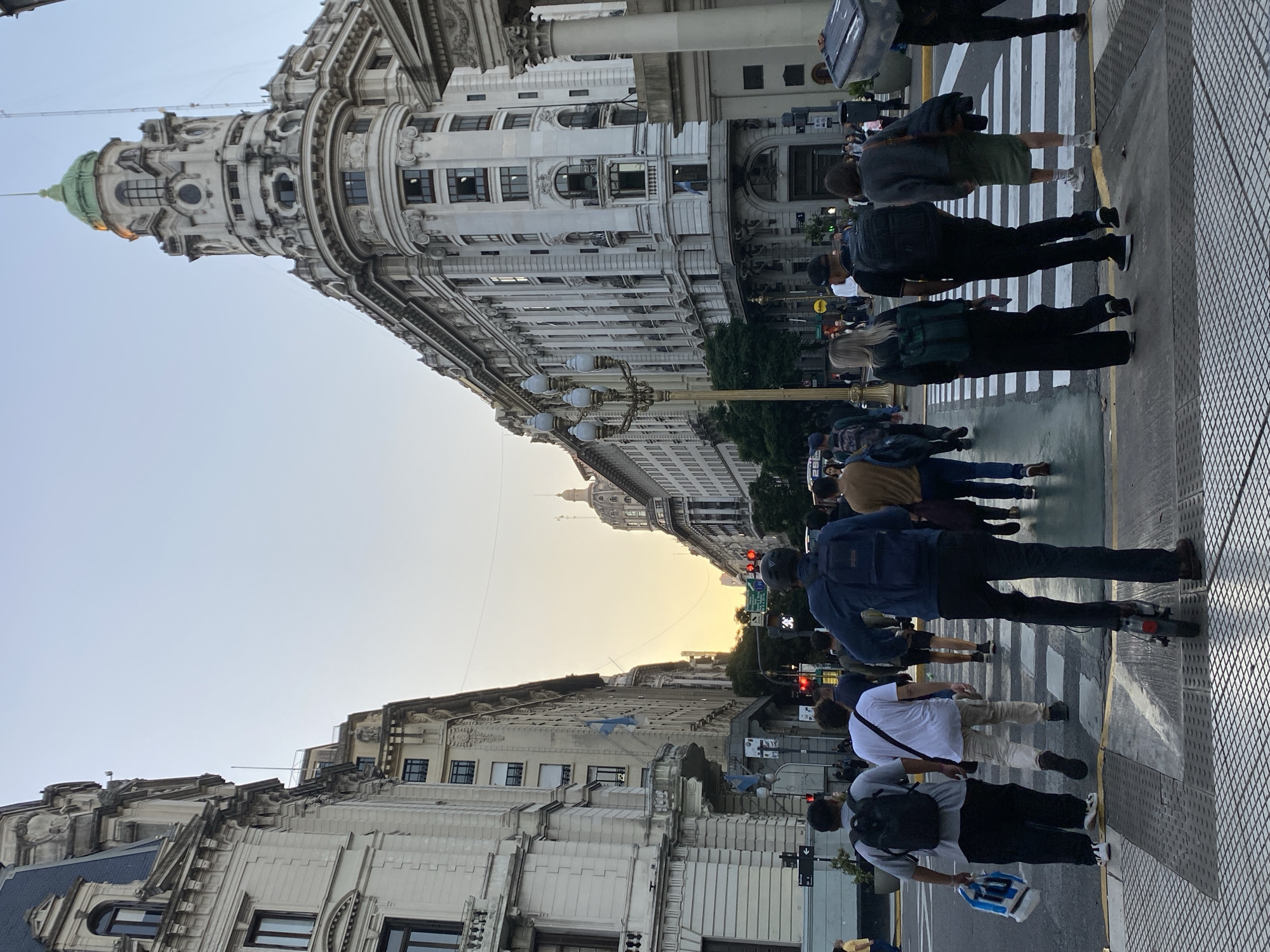 The image shows a street scene in Buenos Aires, Argentina. Pedestrians cross a zebra crossing. The buildings are ornate, with architectural details, including a green-domed building in the background, which is the Palacio de Aguas Corrientes. It appears to be sunset. People are walking, and traffic lights are visible.