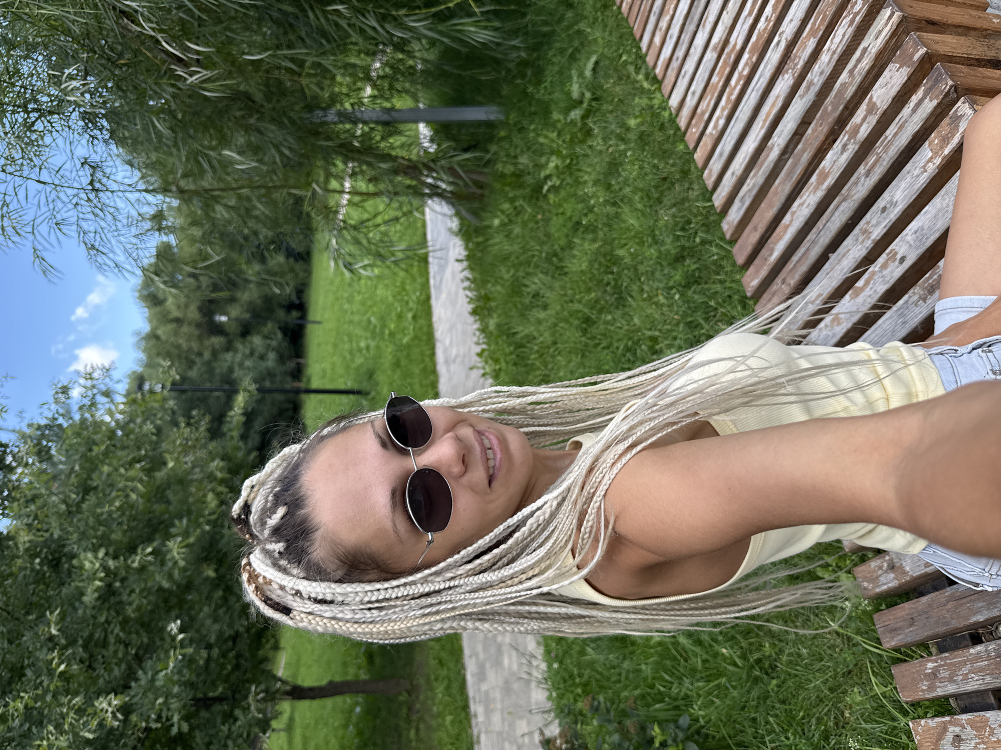 A young woman with blonde braids and sunglasses is sitting on a wooden bench in a park in Rusanovo, Russia. She is wearing a yellow tank top and denim shorts. The park has green grass, trees, and a paved walkway. The weather appears to be sunny. The woman is holding a phone in her right hand and taking a selfie. The image is taken from a slightly elevated angle, looking down at the woman.