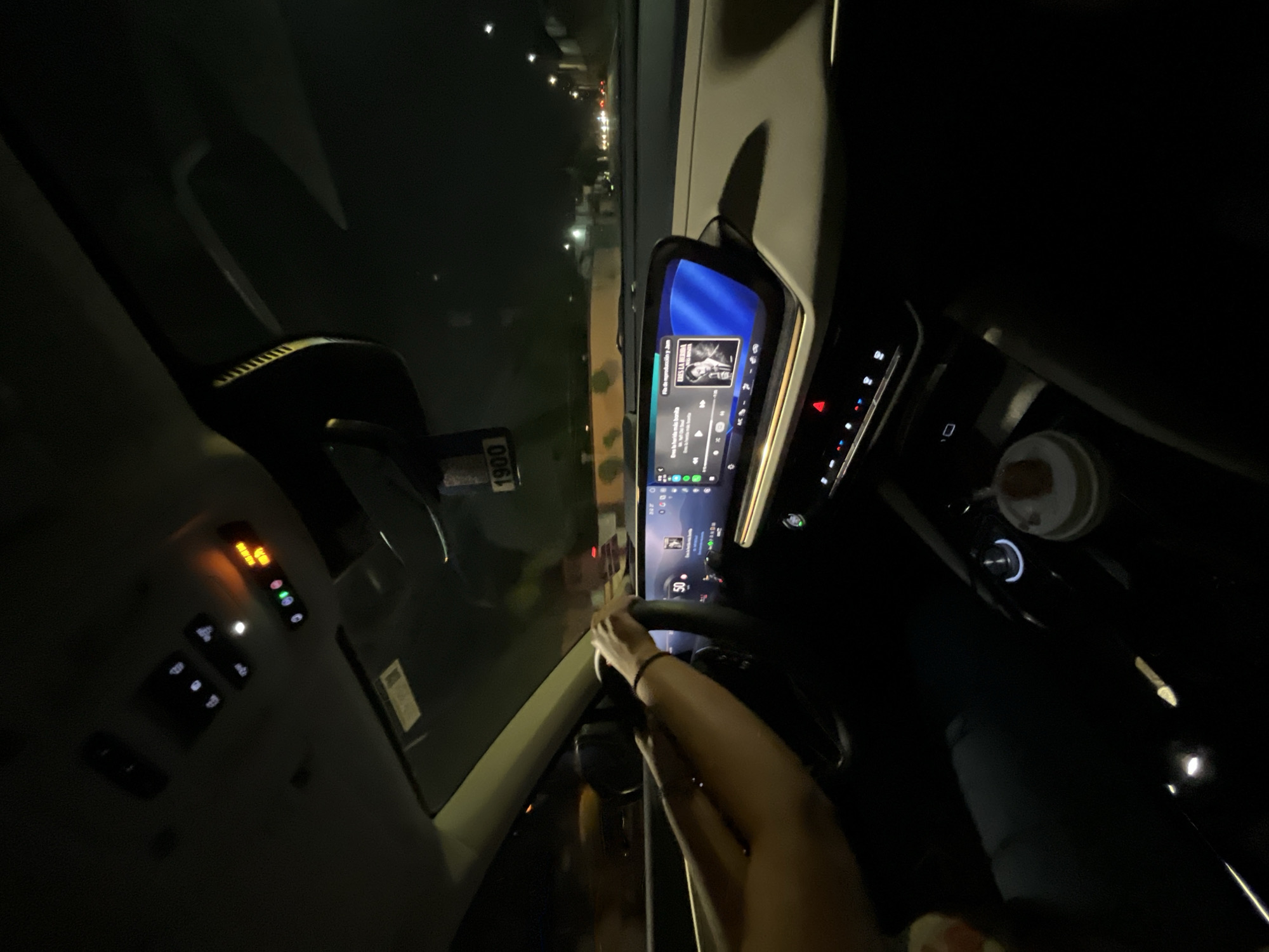 The image shows the interior of a car driving at night in San Bernardino Tlaxcalancingo, Mexico. The dashboard is visible, featuring a large digital display screen playing music. The car's interior lights and exterior street lights create a mix of illumination. A person's arms and hands are visible on the steering wheel. The environment outside is dark, showing glimpses of buildings and street lights. The car appears to be in motion on a road.