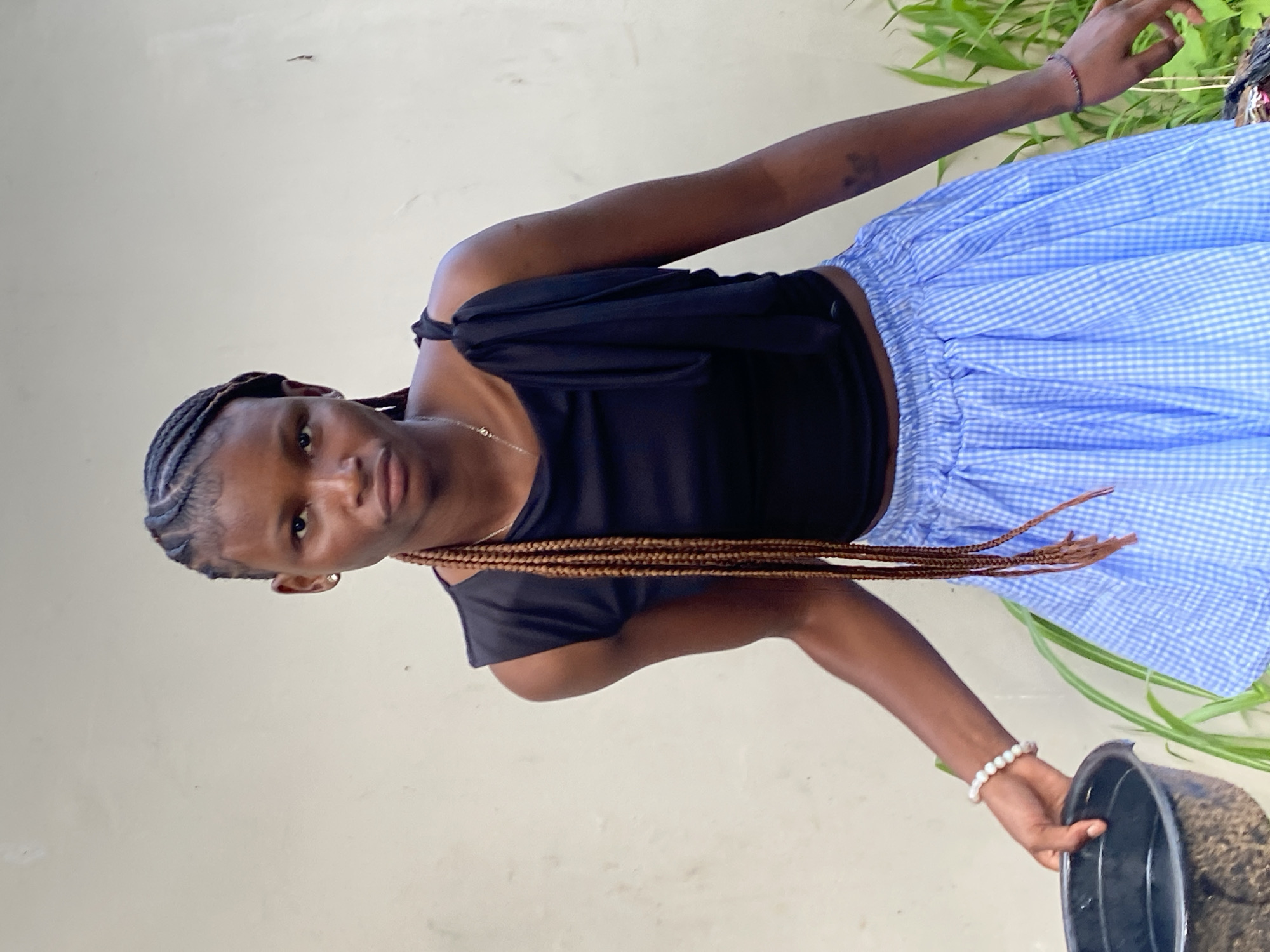 A young woman with braided hair and a black top with a ruffled neckline and a blue and white checkered skirt stands in front of a light-colored wall, holding a black bowl in her left hand. She wears a pearl bracelet on her left wrist and a thin necklace. Green plants are visible to her right. The scene is likely outdoors, possibly in Bar Jirgi Summa, Nigeria, given the context.