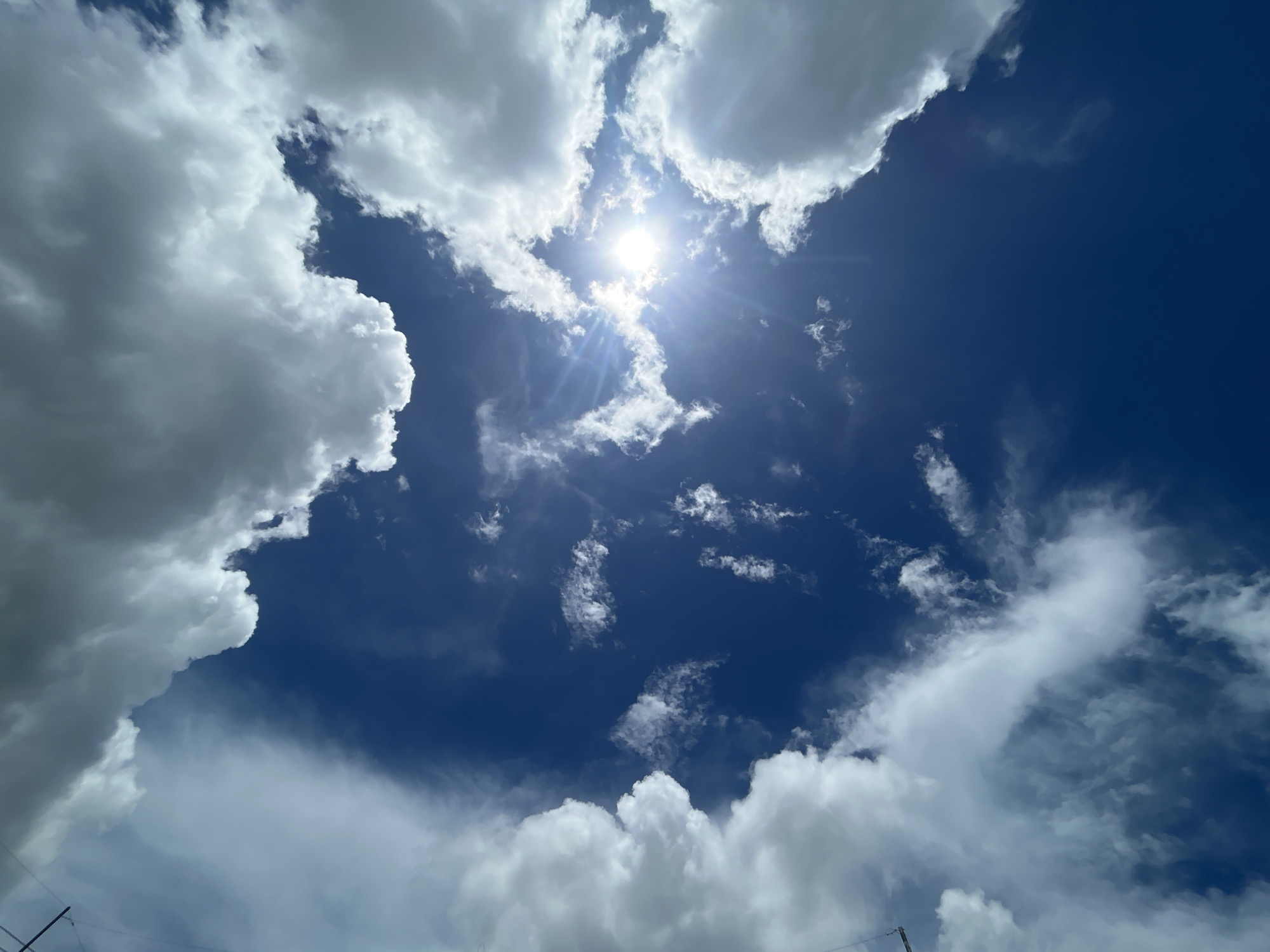 A daytime view of the sky in Baan Hin Kong, Thailand, reveals a bright sun surrounded by scattered clouds. The sky is a deep blue, with large, puffy white clouds framing the central area. Thin, wispy clouds are also visible throughout the sky.