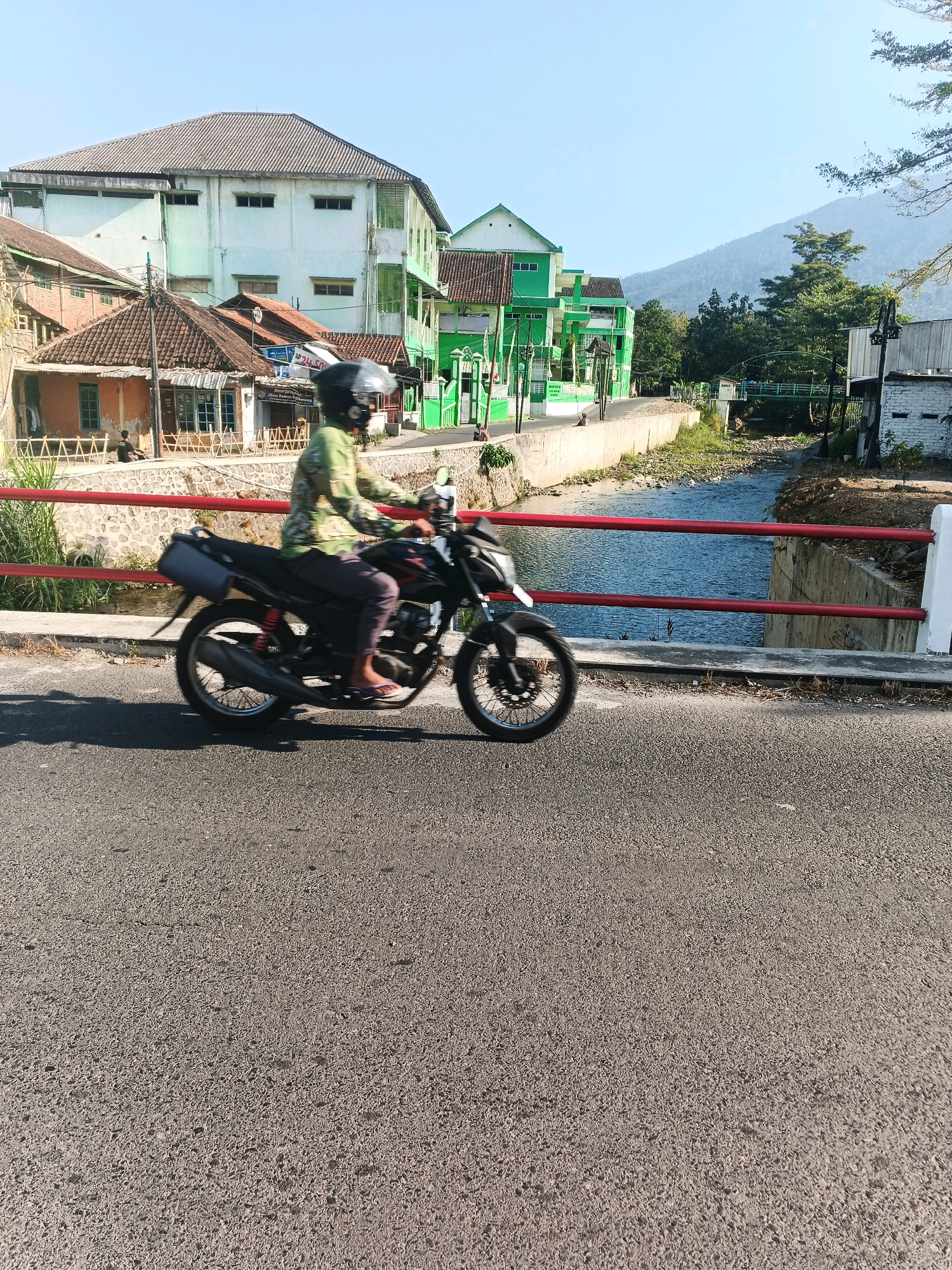 A person wearing a helmet and a patterned shirt is riding a motorcycle across a bridge. The motorcycle is black with red accents. The rider's legs are visible, and they appear to be wearing flip-flops. The bridge has red railings and a concrete barrier. On the left side of the bridge, there are buildings with green and white facades, and houses with brown tile roofs. A river flows beneath the bridge, with vegetation on its banks. In the background, a tree-covered mountain is visible under a clear blue sky. The scene appears to be in a rural or semi-urban area, possibly in Ungaran, Indonesia, given the architectural style and vegetation. The sun is shining, casting shadows on the road.