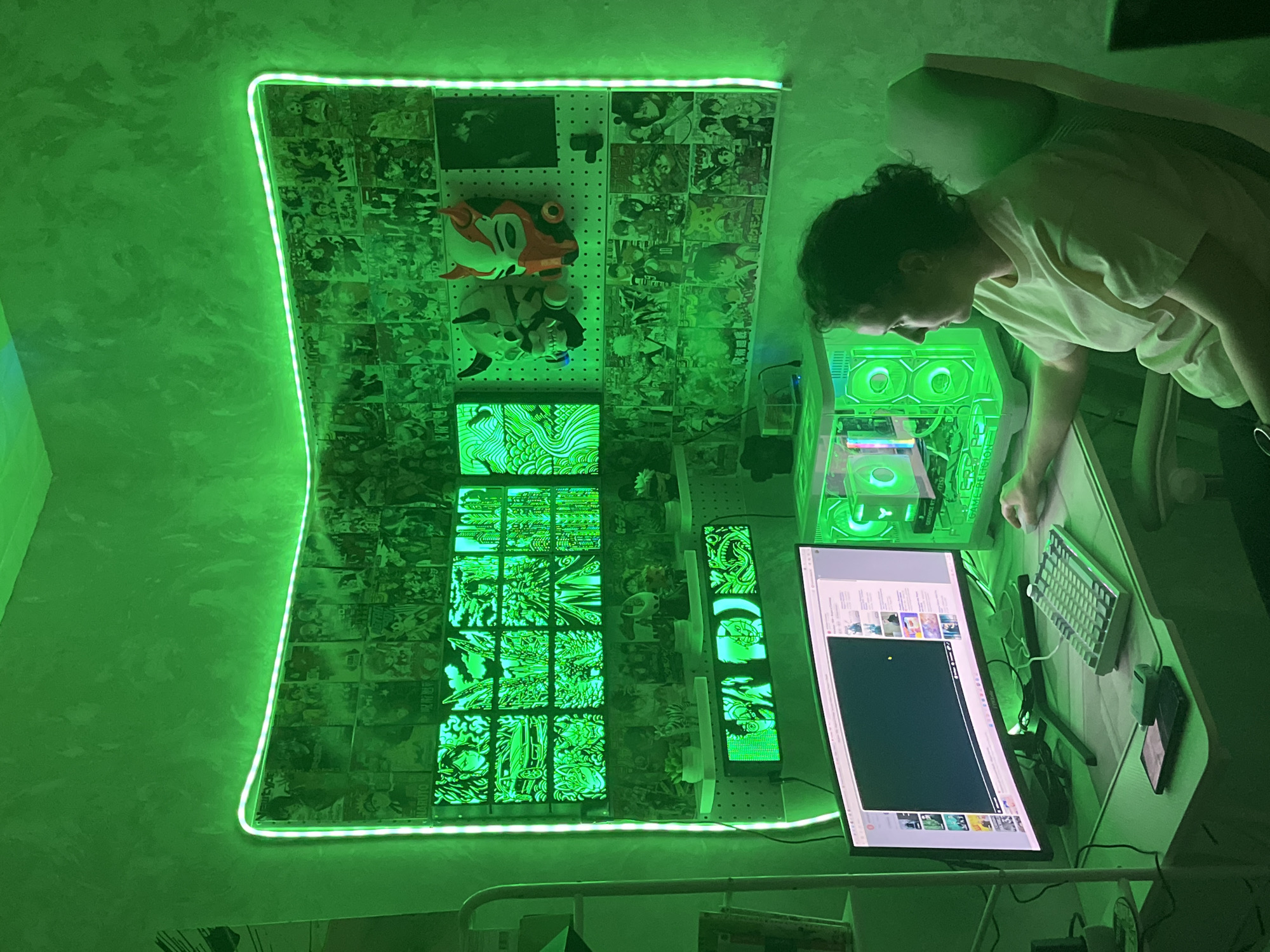 The image shows a person leaning over a desk in a dimly lit room, illuminated by bright green LED lights. The person, with curly dark hair, wears a light-colored t-shirt and is focused on a computer setup. On the desk are a large curved monitor displaying web content, a white keyboard, a phone, and what appears to be a gaming PC tower with internal green lighting.
The walls are textured and painted a dark shade, with a prominent green LED strip tracing the outline of a large display area on the wall. This display area is covered with numerous printed images, possibly from manga or anime, arranged in a grid. Two masks, one red and orange with black details and another white with black accents, hang on a pegboard within this display. Below the main grid of images are several smaller, illuminated panels with intricate green designs.
The overall scene suggests a gaming or creative setup, with a strong emphasis on aesthetics and personalized decor. The green lighting creates an immersive, somewhat futuristic atmosphere. The time of day appears to be evening or night, given the ambient darkness outside the illuminated area. There is no indication of weather or specific location cues beyond the provided context of Moscow, Russia. The person's posture and gaze suggest concentration and engagement with the computer.