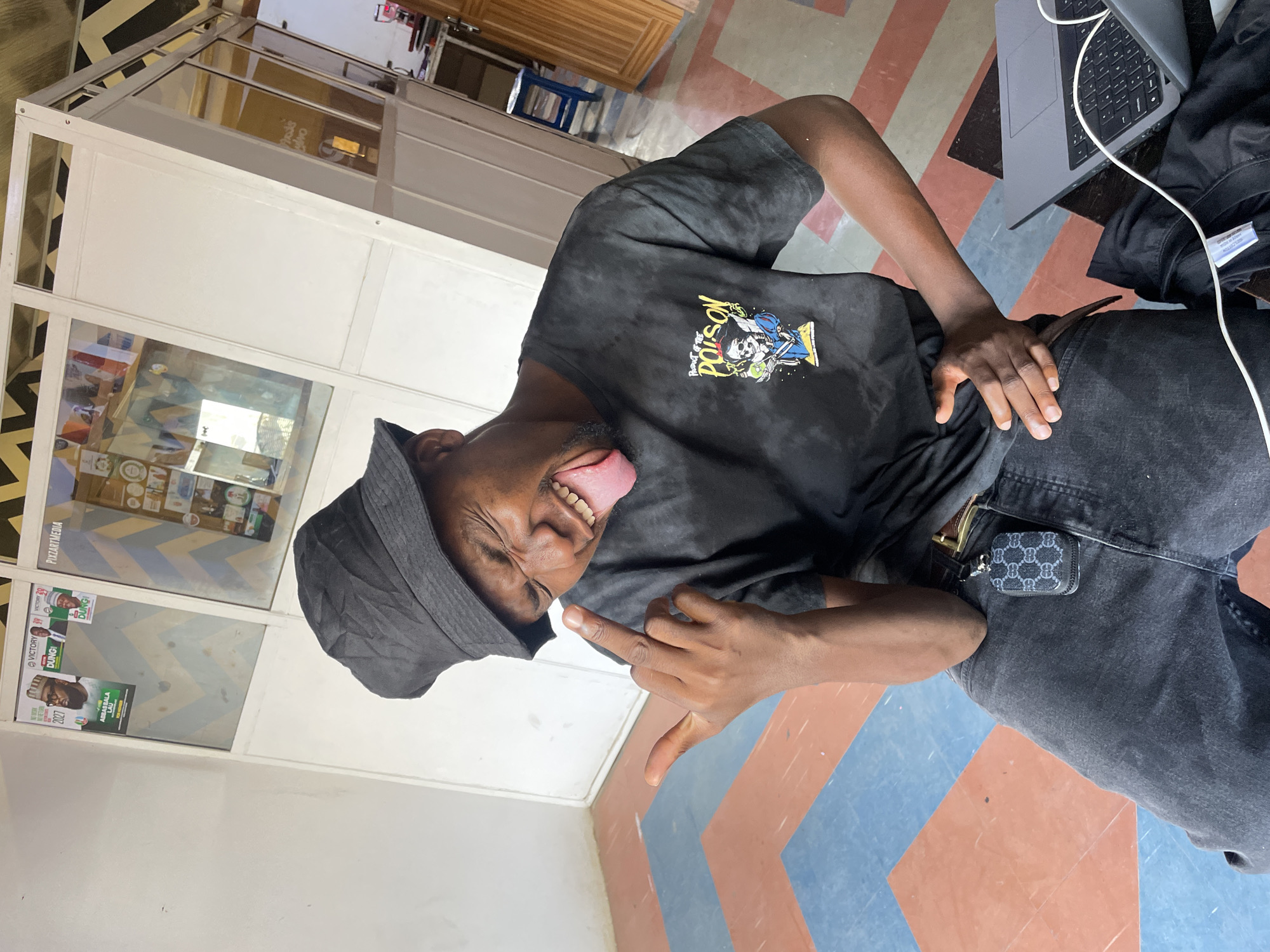 A young man is captured in a playful indoor pose, sticking out his tongue and winking his right eye. He gestures with his left hand, extending his index and pinky fingers, while his right hand rests on his hip. He is dressed in a black tie-dye t-shirt featuring graphic art, including a skull and figures, with the words "POISON" and "PRODUCT OF THE" visible. He also wears black pants, a dark belt with a gold buckle, and a black bucket hat. A small, dark-colored pouch with a repeating interlocking logo pattern is attached to his belt.
The setting appears to be a commercial or institutional building, likely in Jos, Nigeria. The floor features a distinctive geometric pattern in red, blue, black, and white. To the man's left, a glass partition separates him from an inner room. Visible through this partition are several posters, which include political campaign messages such as "VICTORY 20 DUNGI", "DUNGI", and "ABBAS BALA LAU 2027", along with "PIXZARTMEDIA". Inside the room, an office-like environment with shelves and other objects can be discerned. A laptop with a white charging cable is positioned on the floor to the man's right. The lighting suggests it is daytime, and the man's expression conveys a humorous and relaxed demeanor.