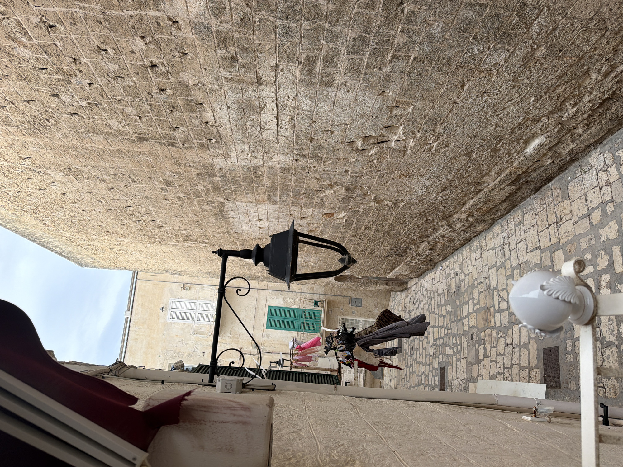 The image depicts a narrow cobblestone alleyway in the historic center of Monopoli, Italy. The left side of the alley is lined with a pale yellow building, featuring a red awning overhead. A white pipe runs vertically along this building's exterior. Clothes are hung out to dry on a line strung across the alley, with various garments including a pink dress and grey shirts visible. A traditional black street lamp with decorative scrollwork is mounted on the wall. The right side of the alley is dominated by a thick, ancient stone wall constructed from irregular, rough-hewn blocks. The wall exhibits signs of age and weathering, with visible erosion and indentations. In the background, a building with green shutters and a white-railed balcony can be seen. The ground is paved with worn cobblestones. The overall atmosphere suggests a typical Italian coastal town scene, characterized by old architecture and everyday life.
