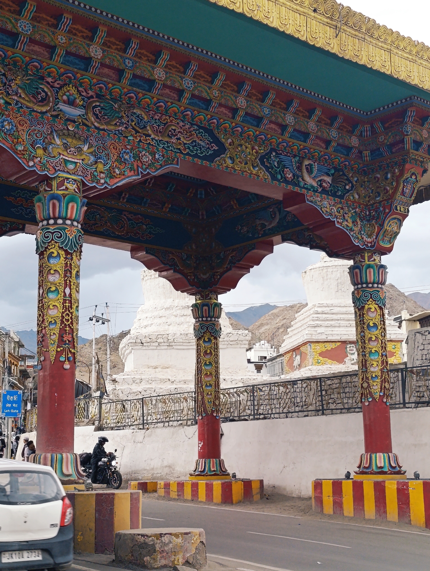 The image depicts a view of the Leh Gate, also known as the "Leh Gateway," in Leh, India. The gate is an ornate structure featuring vibrant colors and intricate carvings. Two large red pillars, adorned with intricate golden and colorful patterns, support a richly decorated roof. The roof itself is a marvel of craftsmanship, with detailed depictions of mythical creatures like dragons and birds, along with floral motifs.
In the background, several white stupas, a common architectural feature in Buddhist regions, are visible. The stupas are large, domed structures, with one prominently positioned behind the gate. Beyond the stupas, the arid, mountainous landscape characteristic of Leh is apparent under a cloudy sky.
In the foreground, a motorcycle rider, wearing a helmet and black jacket, is stopped at the gate. A white car is partially visible in the lower left corner of the image. The road in front of the gate is marked with yellow and red striped barriers. A blue sign with Tibetan script and the English words "Leh Gate" is also visible on the left side of the image, indicating the location. The scene is set on a street, suggesting an entrance or significant point within the city.