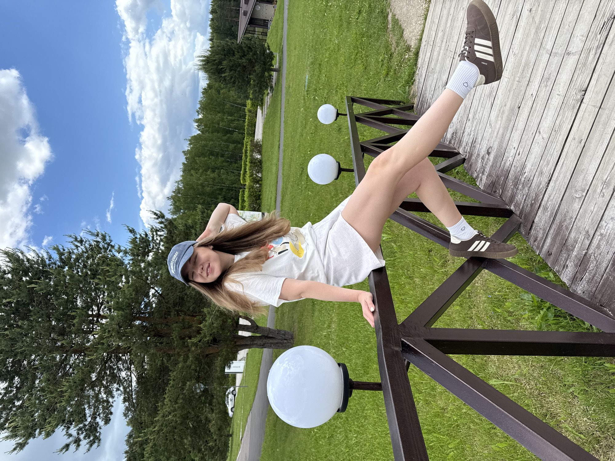 A young woman is captured lounging outdoors on a bright, sunny day in Dobrograd, Russia. She is lying sideways on a dark brown metal railing, which supports a wooden walkway or bridge, with her head turned to face the camera and a slight, relaxed smile. One leg is bent and resting on the railing, while the other is extended upwards.
She is dressed in a casual outfit consisting of a white t-shirt with a partial graphic design, light-colored shorts or a skirt, and a light blue baseball cap that reads "SOUND COAST". Her feet are adorned with light blue ankle socks featuring small heart patterns and dark brown Adidas Samba sneakers with white stripes, revealing the "SAMBA" branding on the side.
The scene is set in a verdant outdoor environment, likely a park or resort. The background showcases expansive green lawns, numerous trees, and a paved path or road. A modern building with a dark roof is visible in the distance, nestled among the foliage. Several white, globe-shaped lights on dark posts are integrated into the railing structure in the foreground. The sky is a clear blue with scattered white clouds, indicating pleasant weather conditions during daylight hours.