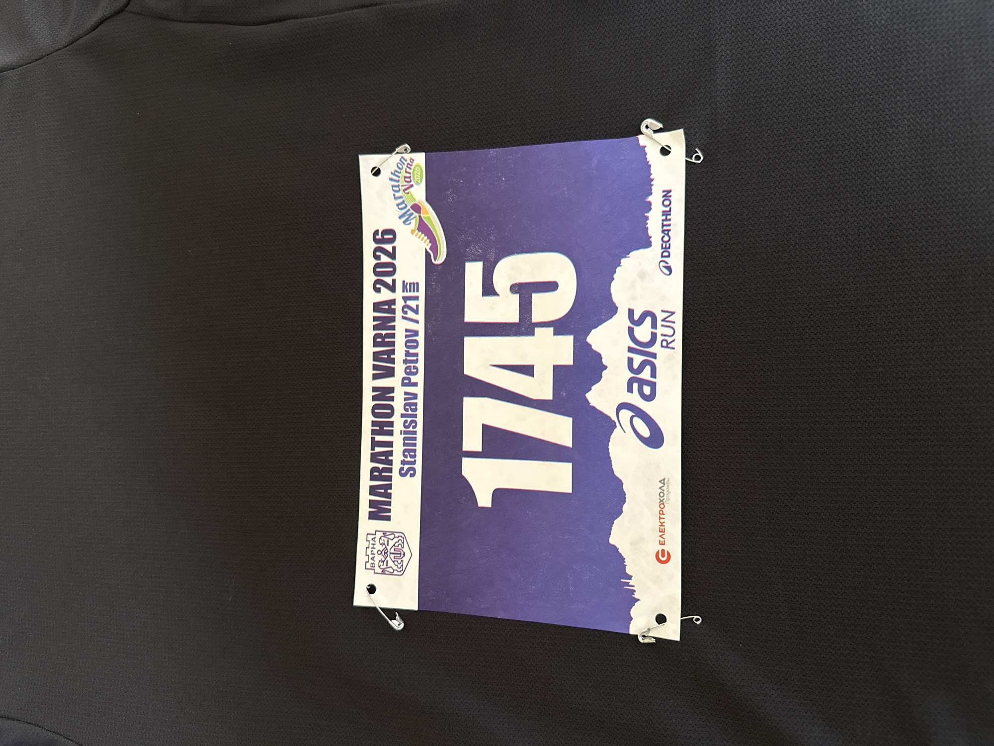 The image shows a runner's bib attached to a black running shirt with safety pins. The bib is for the Marathon Varna 2026. The name "Stanislav Petrov" is printed on the bib, along with the race number "1745". Logos for "asics RUN", "DECATHLON", and "ЕЛЕКТРОХОЛД" (Elektrohold) are also present on the bib. A colorful logo depicting a running shoe is featured, with the text "Marathon Varna 2026" below it. The shirt appears to be made of a synthetic, possibly moisture-wicking, material.