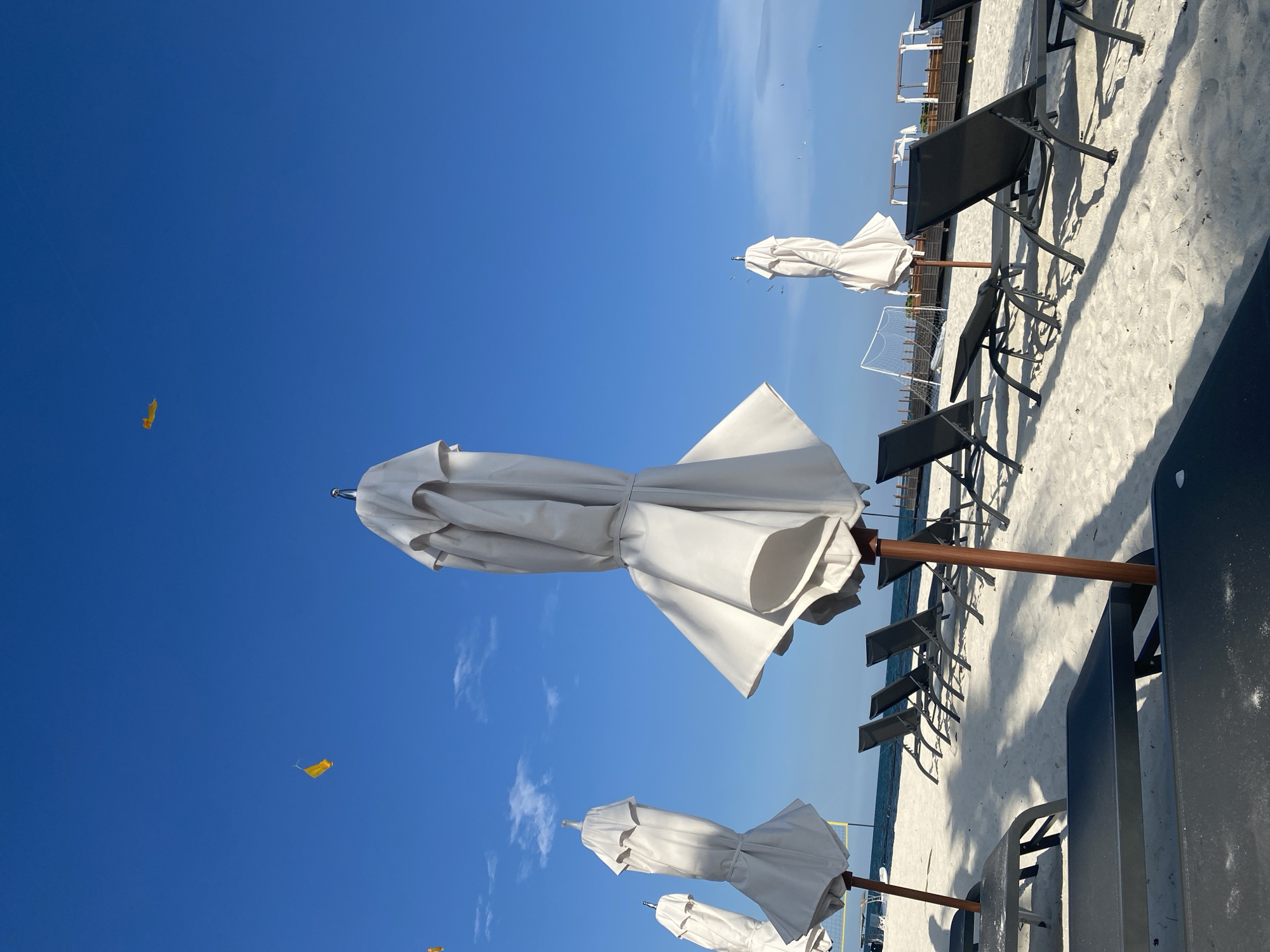 The image shows a beach scene in Marina Maroma, Mexico. The view is oriented towards the sky, which is a clear blue with scattered white clouds. Several closed white beach umbrellas are positioned at varying distances, interspersed among dark beach chairs on a sandy surface. In the distance, a soccer goal is visible on the beach. Some small yellow kites are scattered in the sky. The overall composition is bright and depicts a typical beach environment during daytime.
