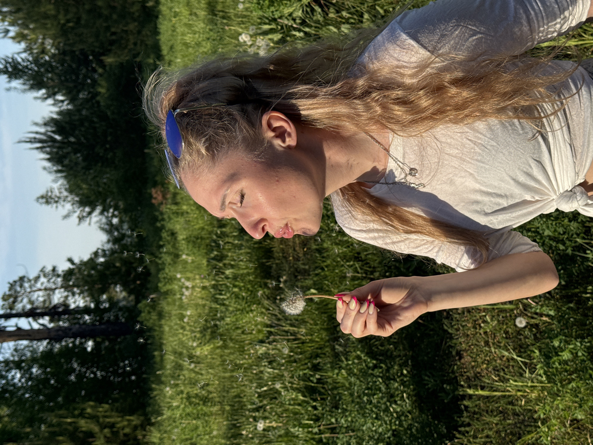 A young woman with long, blonde hair is blowing on a dandelion in a grassy field. She is wearing a light-colored shirt and has blue sunglasses perched on her head. Some dandelion seeds are floating away from the flower. A line of trees is visible in the background. While the prompt asks for the location in Прикамский, Russia, this cannot be determined from the image.