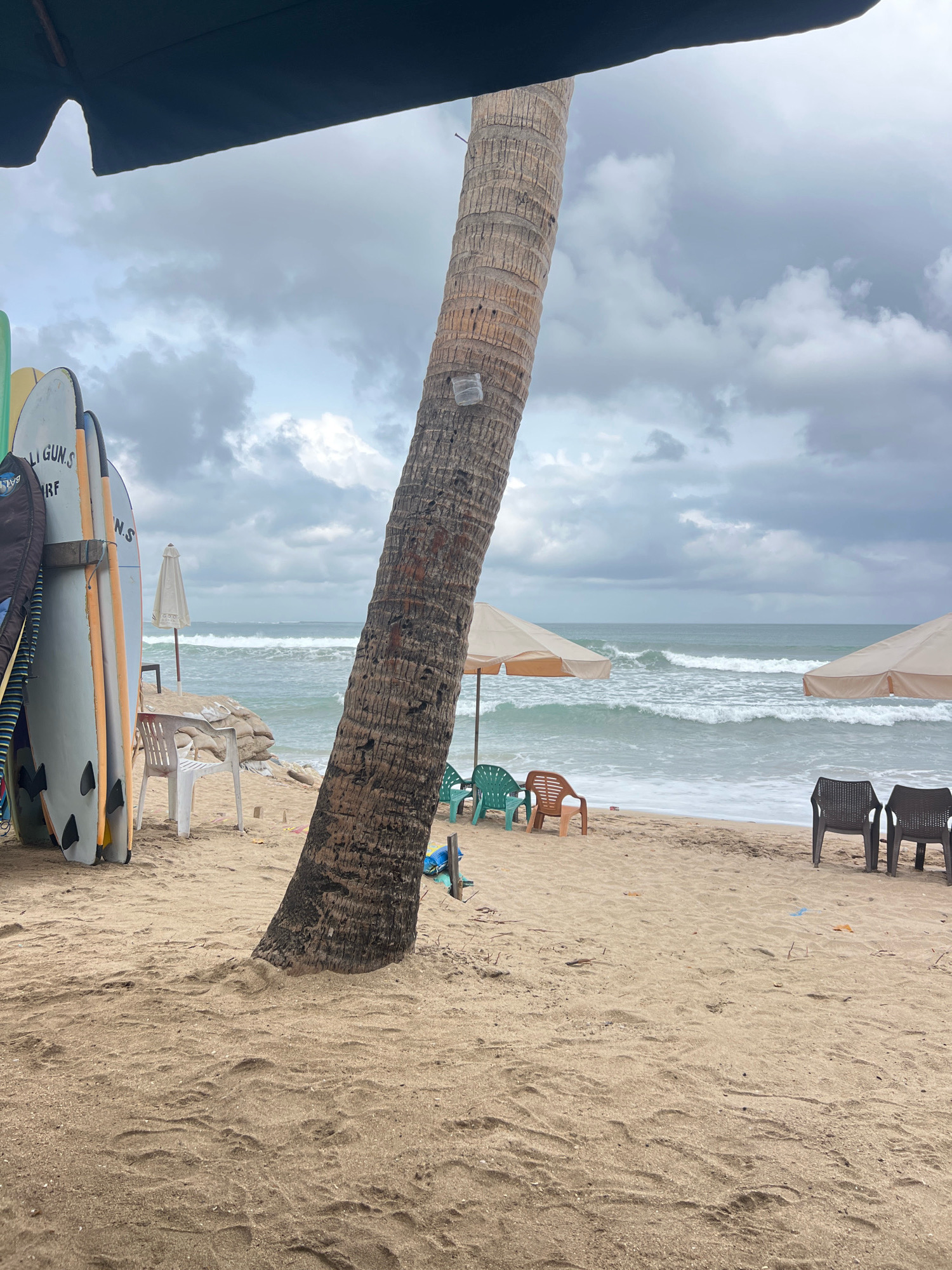 The image depicts a beach scene in Kuta, Indonesia, on an overcast day. In the foreground, sand covers the ground, with footprints visible. A large palm tree trunk stands prominently in the center, leaning towards the right. To the left of the palm tree, several surfboards are propped up, leaning against each other. Behind the surfboards, a white plastic chair sits on the sand. Further back, a light-colored beach umbrella is open, positioned near a rocky embankment. The ocean stretches out to the horizon, with moderate waves breaking on the shore. In the midground, two more beach umbrellas, one on the left and one on the right, are set up, each with chairs placed in front of them. The umbrella on the left has two green plastic chairs and one brown plastic chair in front of it. The umbrella on the right has two dark-colored chairs. The sky is filled with heavy, grey clouds.