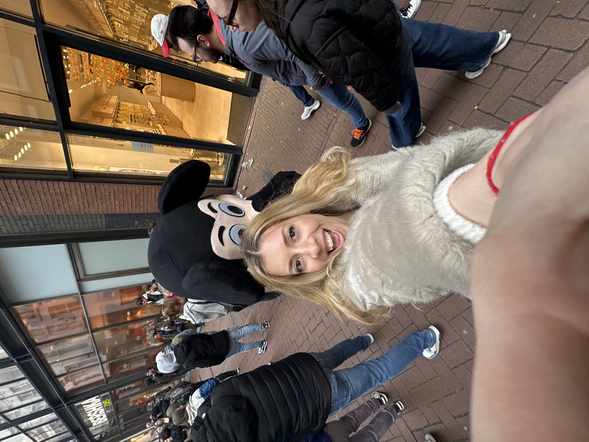 A distorted, wide-angle, low-angle selfie captures a woman with long blonde hair, wearing a fuzzy white sweater, smiling while posing with a Mickey Mouse mascot on a brick sidewalk, with a group of people in the background. The photo appears to be taken on a commercial street in Amsterdam, Netherlands, as indicated by the storefronts and architecture. Several people are visible, standing and walking along the sidewalk, some wearing jackets and jeans. A glass-fronted store with displays inside is visible to the left of the image.
