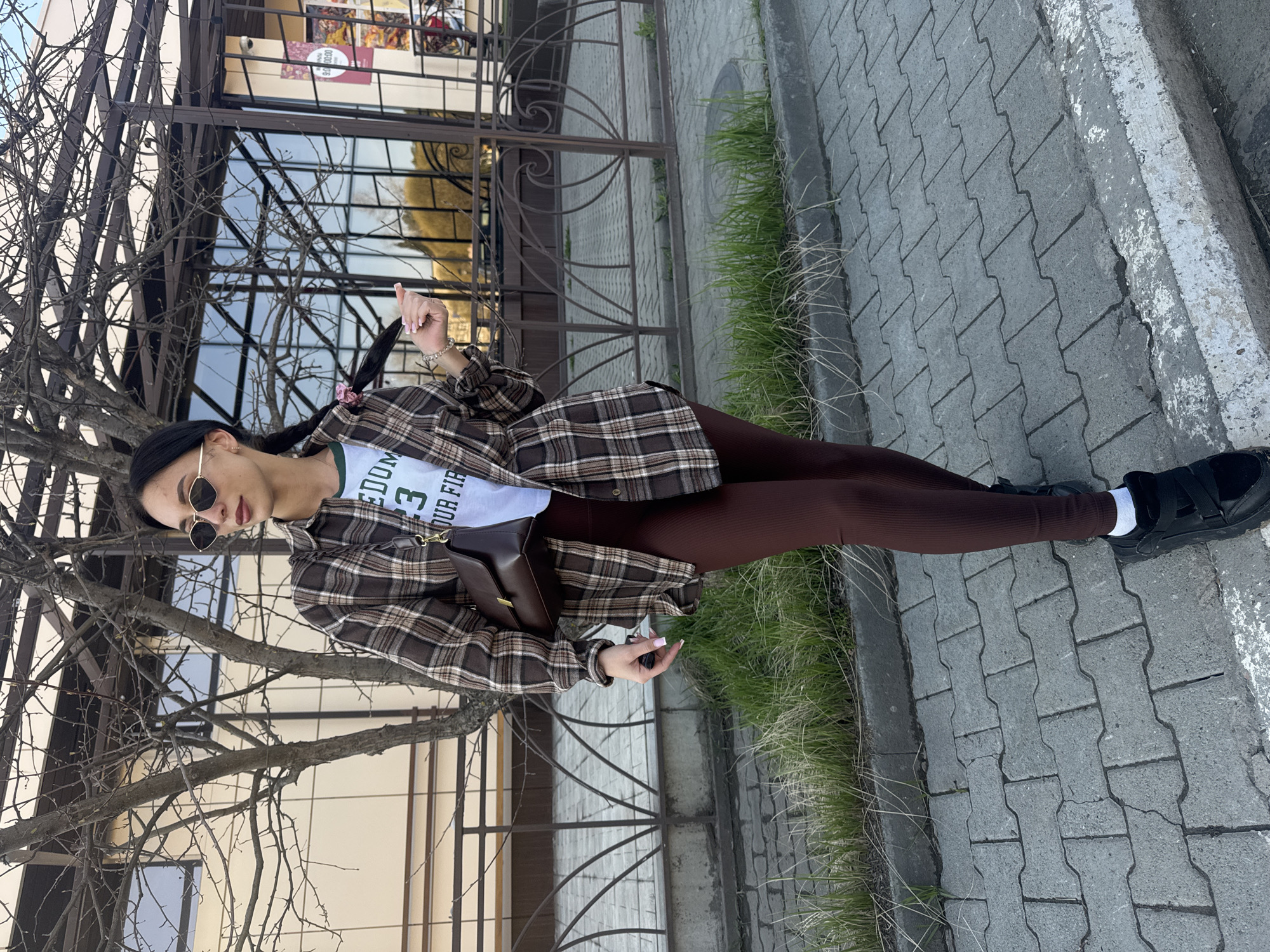A woman stands on a sidewalk in Chaikovsky, Russia. She is wearing sunglasses, a plaid jacket, brown leggings, and black shoes with white socks. She has a brown purse slung over her shoulder. In her hand, she holds an object that appears to be keys or a small electronic device. Behind her is a metal fence and a building with windows. A tree with bare branches is visible in the foreground. The sidewalk is made of interlocking gray pavers. Grass is growing along the edge of the sidewalk.