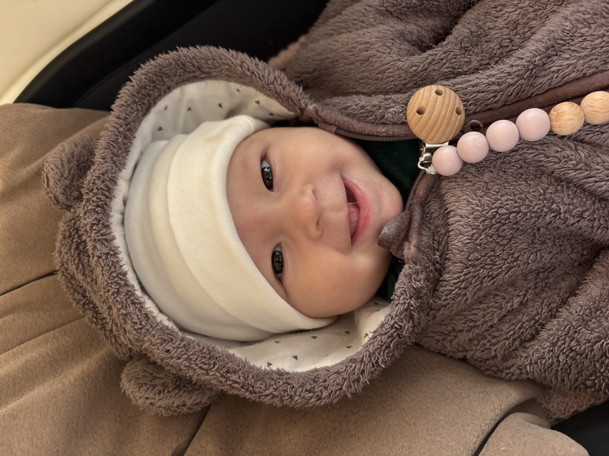 A baby wearing a white cap and brown furry hooded jacket lies on a brown surface. A pacifier clip with light pink and wooden beads is attached to the jacket. The location is not identifiable as Sochi, Russia.