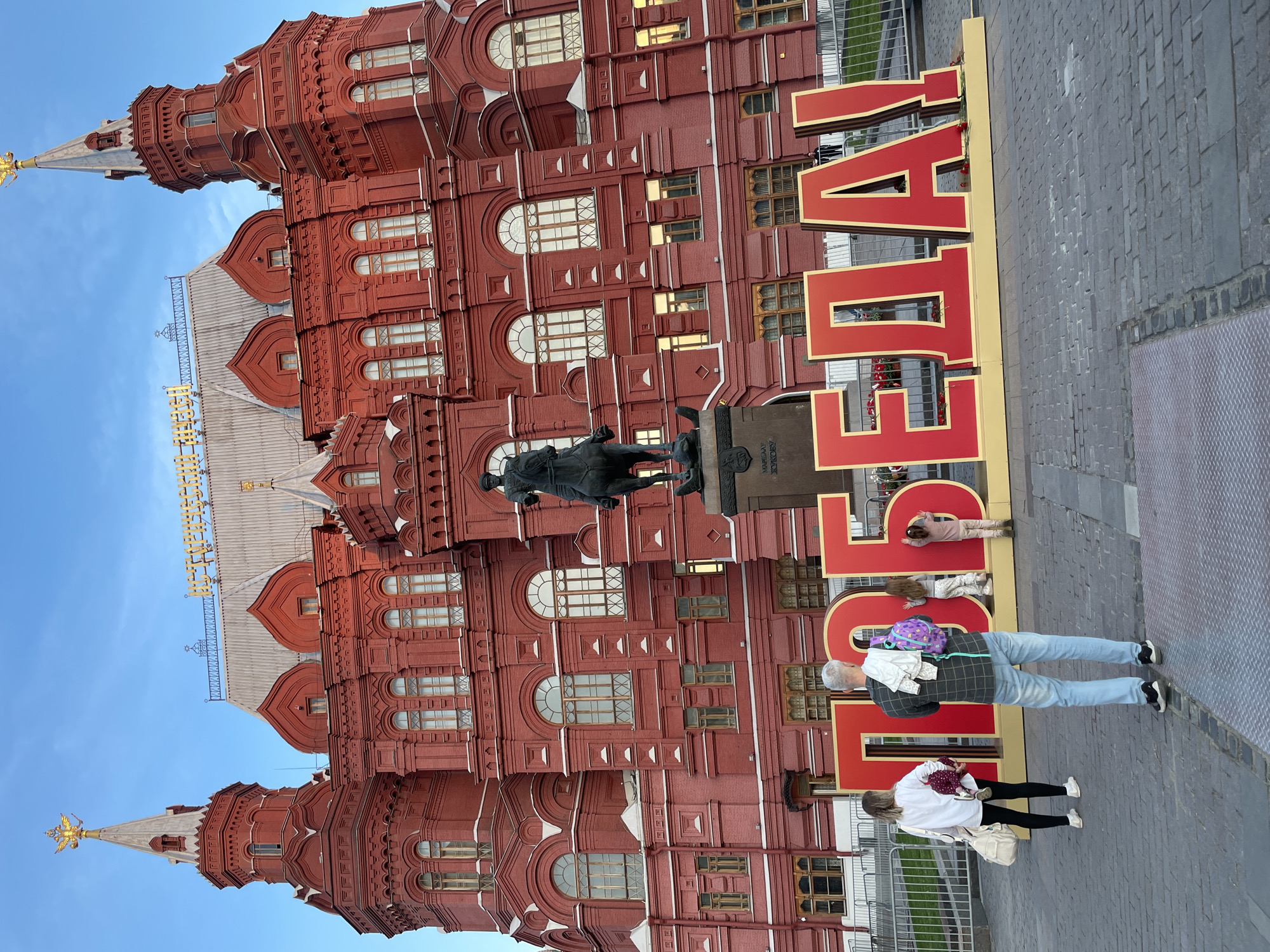 A medium shot depicts the State Historical Museum on Red Square in Moscow. The building is large, red, and ornate. In front of the museum, there is a bronze equestrian statue on a stone pedestal. The word "ПОБЕДА", the Russian word for "victory", is displayed in large red letters with gold trim. The text is arranged vertically in the center foreground. The letters are so large that they partially obscure the view of the building. Several people are standing and interacting with the sign. A person in blue jeans and a gray plaid shirt is standing in front of the letter "Д". There is a white garment hanging on their back and a purple backpack lying on the letter. A person in a white shirt and black leggings is standing on the right side of the letter "О". Two children are lying on the letter "Б". The sky is blue.