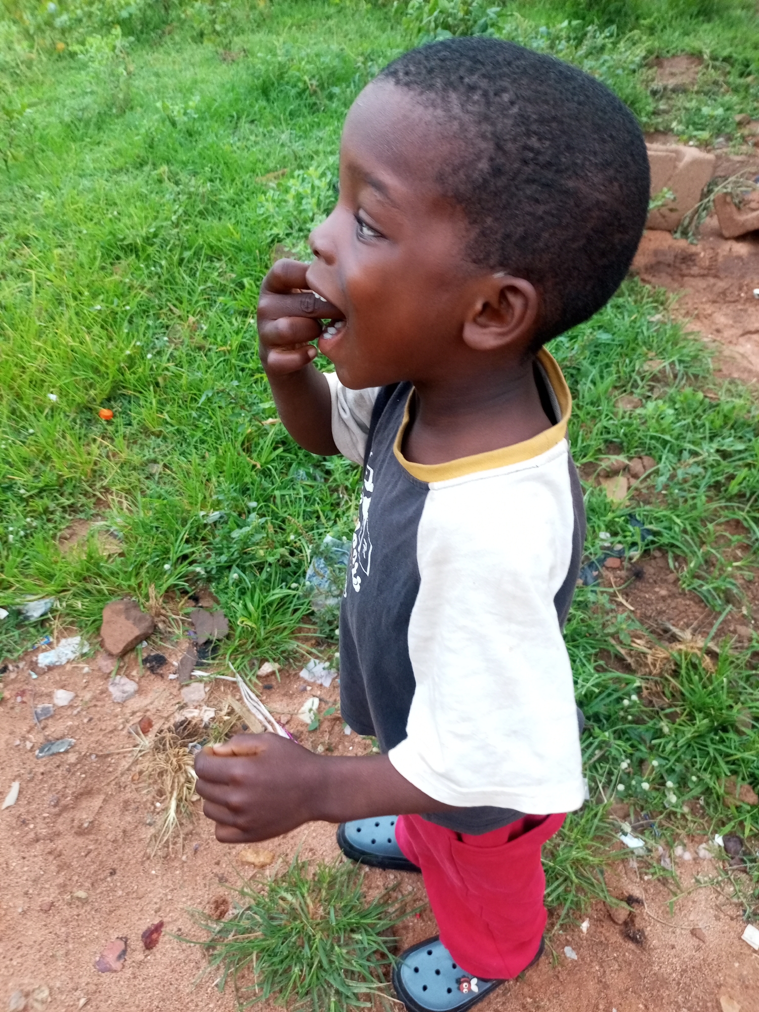 A child, appearing to be of African descent, is captured in a medium shot, facing to the left in profile. The child has dark skin, short black hair, and is wearing a t-shirt with a black upper section and white sleeves, accented with a yellow trim around the collar. Their lower body is clad in red pants. The child's right hand is raised to their mouth, and they appear to be putting something in their mouth. They are standing on a sandy, dirt ground with patches of green grass and scattered debris, suggesting an outdoor, possibly urban or suburban, environment. In the background, more green grass and some brown earthy areas are visible. The lighting suggests it is daytime.