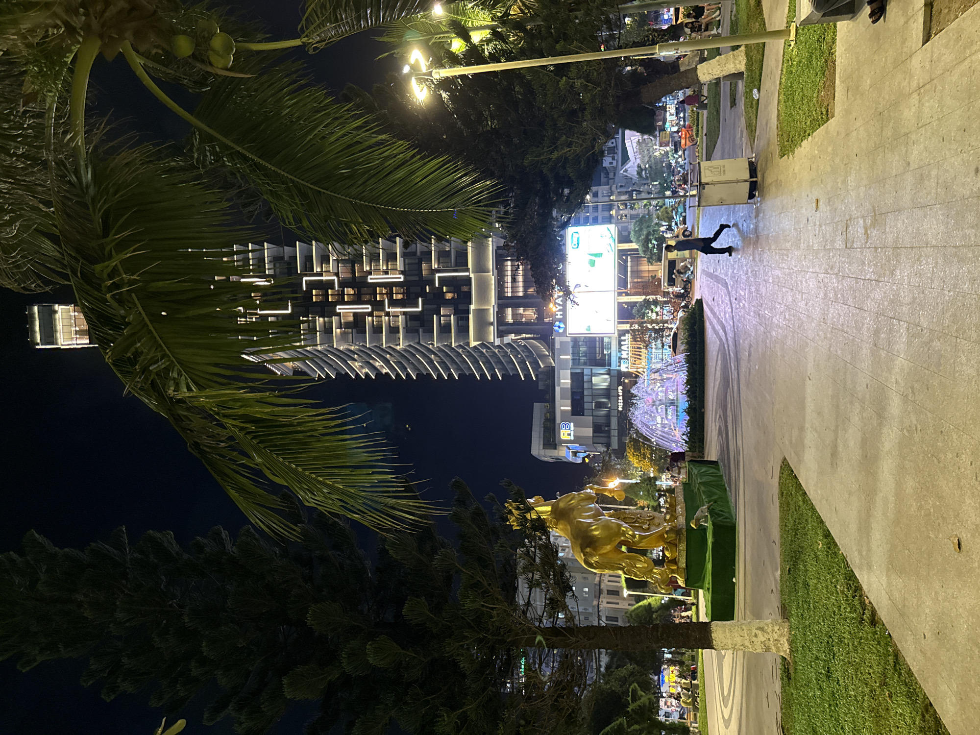 At night, in Nha Trang, Vietnam, a golden horse statue is displayed in a park area. Behind the statue, a modern skyscraper with illuminated windows dominates the skyline. A colorful fountain adds to the ambiance. Palm trees and other vegetation are visible, creating a tropical atmosphere. People are walking in the park, and vehicles are present on the street in the background, indicating a busy urban setting. The scene is well-lit by streetlights and the building's illumination, contributing to a lively night view.
