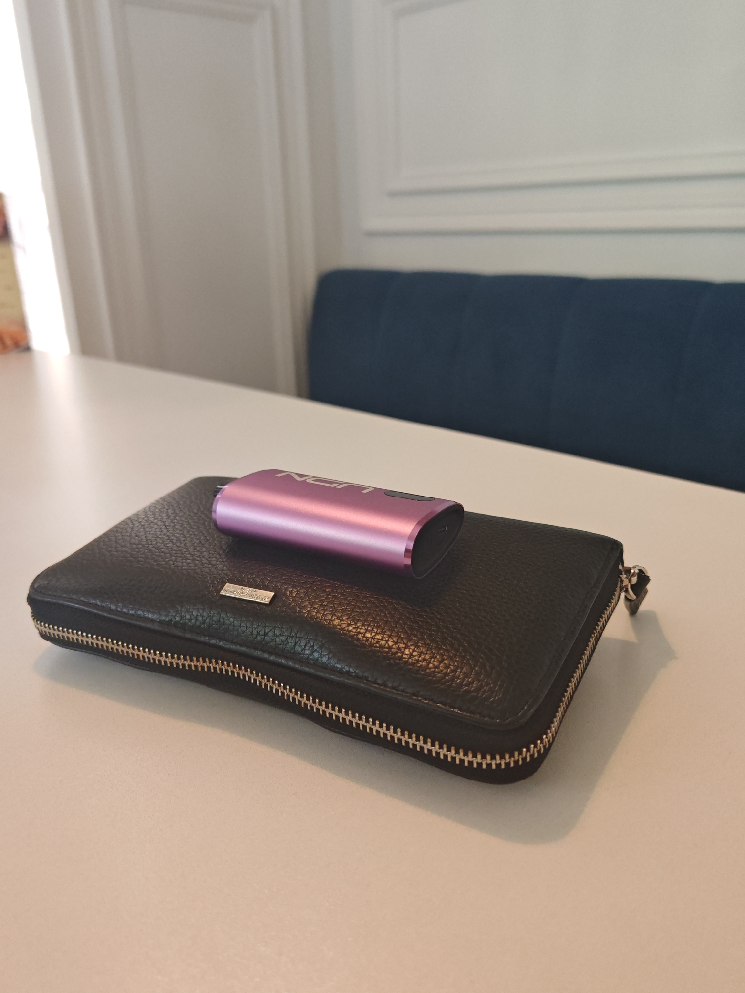 A close-up, slightly elevated shot captures a black leather wallet and a metallic purple electronic device resting on a white table. The wallet, horizontally oriented, features a textured surface, a visible zipper running along three sides, and a small, silver-colored rectangular plaque with an unidentifiable logo on its front. Perched centrally on the wallet is the sleek, cylindrical purple device, which has "IQOS" faintly visible in white lettering on its side.
The background reveals a clean, modern indoor setting. A bright light source, likely a window, is positioned to the left, casting soft illumination across the table. Behind the table, a white wall with decorative molding extends upwards. To the right, a segment of a dark blue, possibly velvet or upholstered, bench or sofa is visible, providing a contrasting color against the otherwise light-toned environment. No specific activity is taking place; the items are simply at rest. The lighting suggests it is daytime. The image lacks any explicit cues to the specified location of Sochi, Russia, beyond being an interior space. No people are clearly visible, and no particular emotion is conveyed.