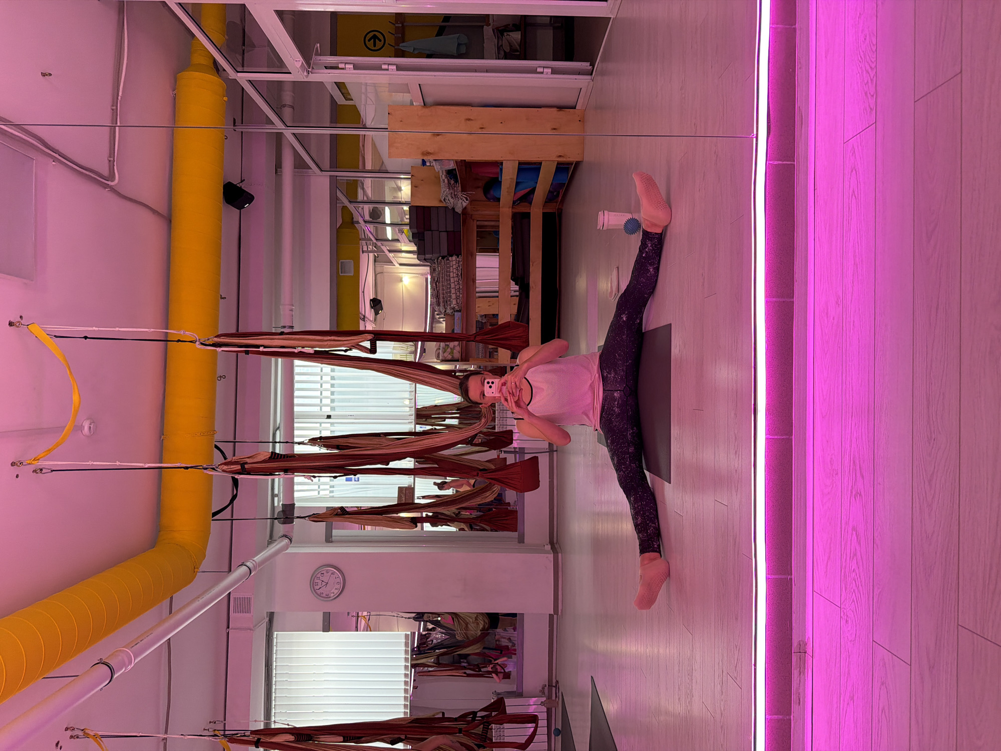 A woman is taking a selfie in a yoga studio. She is sitting on a yoga mat in a straddle split position, facing the mirror. She is wearing a pink tank top and black patterned leggings. Her phone is held up in front of her face, and she is looking at the screen. The studio has aerial yoga hammocks hanging from the ceiling, mirrors on the walls, and wooden floors. There is a clock on the wall that reads 10:25. The lighting in the studio is pink. The location is Chaikovsky, Russia.