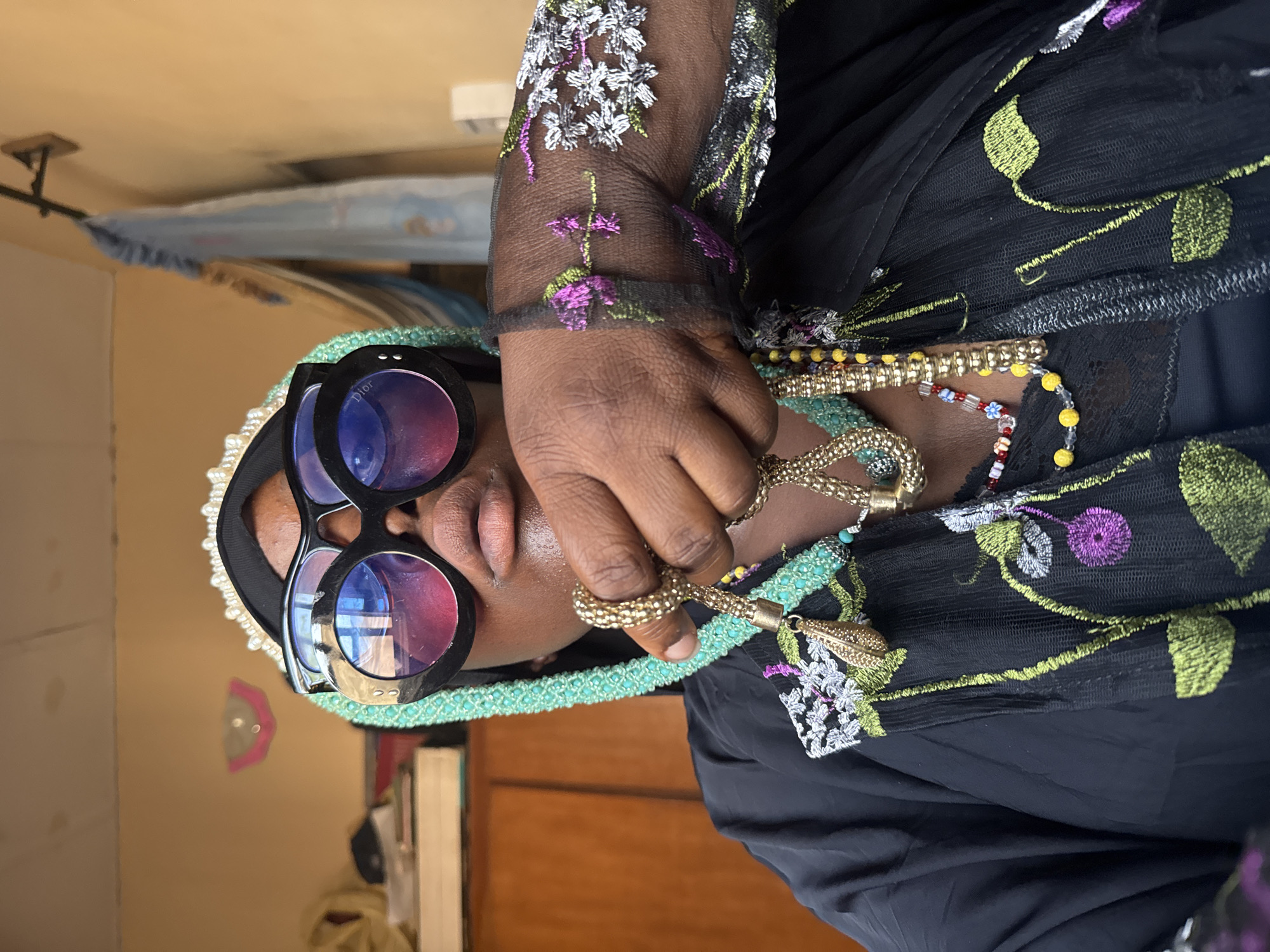 The image depicts a person with dark skin wearing large, round black sunglasses, a head covering with light blue trim and pearl accents, and a black garment with floral embroidery. The person is holding a gold-colored necklace. The background includes a light brown wall and a wooden cabinet. There is no conclusive evidence to determine if this image was taken in Madangala, Nigeria. The image provides insufficient information to verify specific location.