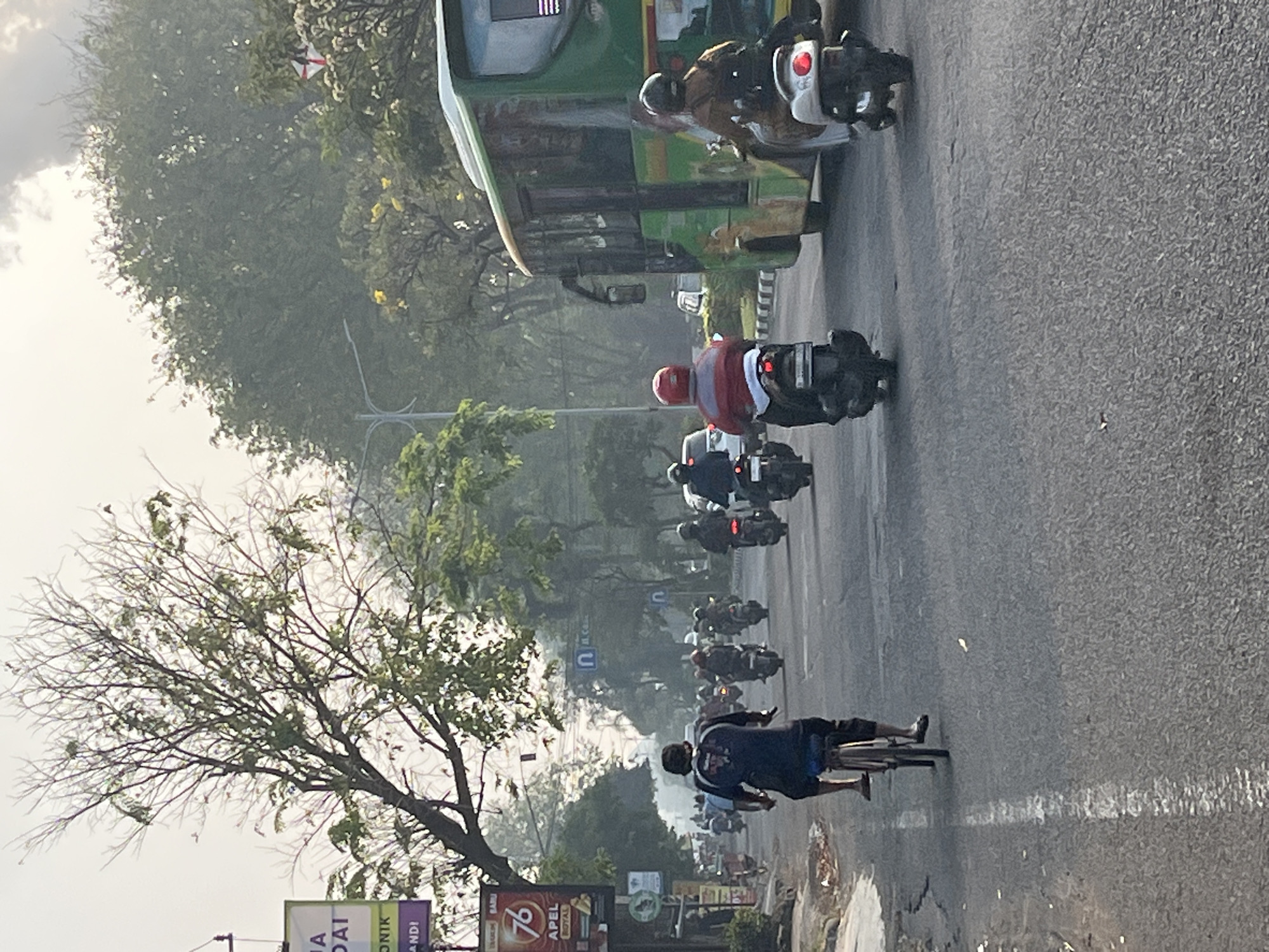 In Candi, Indonesia, a street scene depicts moderate traffic flow. In the foreground, a person on a bicycle rides away from the camera, facing traffic. They are wearing a dark blue long-sleeved shirt and dark pants, with their feet on the pedals. To the right of the cyclist, a scooter with a rider wearing a red and white jacket is visible, also heading away from the camera. Further ahead on the road, multiple other scooters and motorcycles are in motion, forming a line of traffic. A large green bus with yellow accents is positioned on the right side of the road, also moving forward. The road is paved and has a white line marking the lane. On the left side of the road, a billboard with the number "76" and text in Indonesian is visible, along with trees and buildings. The sky is overcast with some visible clouds, and the overall lighting suggests it is either early morning or late afternoon, with a slight haze in the air. The environment appears to be an urban or suburban setting.