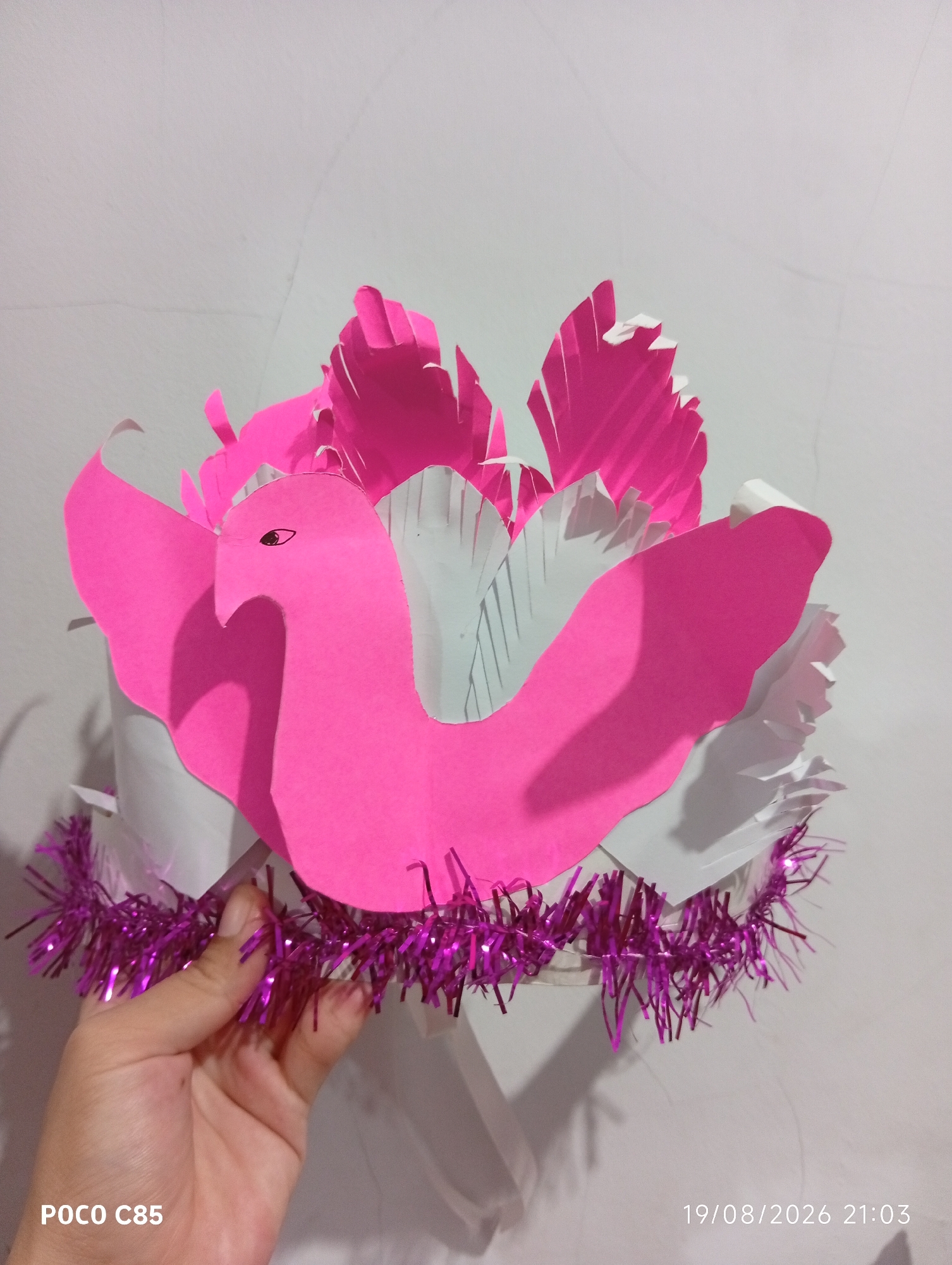 An overhead, eye-level shot shows a pink paper-cutout of a bird on top of a white paper crown adorned with purple tinsel. The crown is held by a hand in the lower left corner of the frame, which is partially cropped out. The bird cutout is in the shape of a flamingo, with a long, curved neck, a beaked head, and wide wings. The bird is positioned on the front of the crown and is facing left, with its eye visible. Behind the bird, several white and pink paper cutouts resembling feathers or flames are layered, creating a voluminous effect. The crown itself appears to be made of white paper, and its base is wrapped with a string of shimmering purple tinsel. The background is a plain, light-colored wall, with some faint shadows suggesting the presence of light sources. The timestamp "19/08/2026 21:03" and the text "POCO C85" are visible in the bottom right and left corners of the image, respectively, indicating the time and the device used for capture.