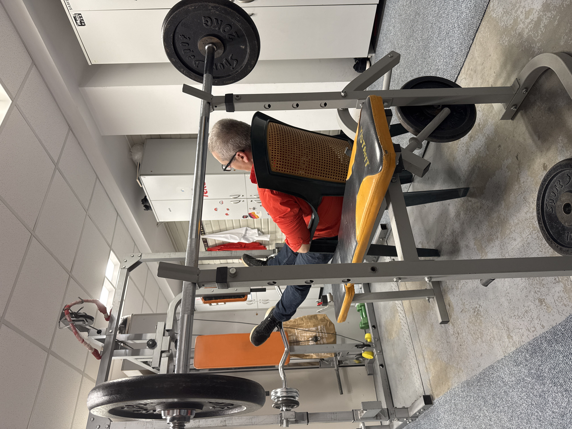 An adult male wearing a red shirt, blue jeans, and black shoes is sitting on a weight bench in a home gym. He is wearing glasses and appears to be looking down. The weight bench is part of a larger weightlifting machine. The gym is located in a room with white walls and a gray carpet. There are various weight plates and other gym equipment visible in the background. The location in the city of Sofia, Bulgaria, cannot be determined from the image.