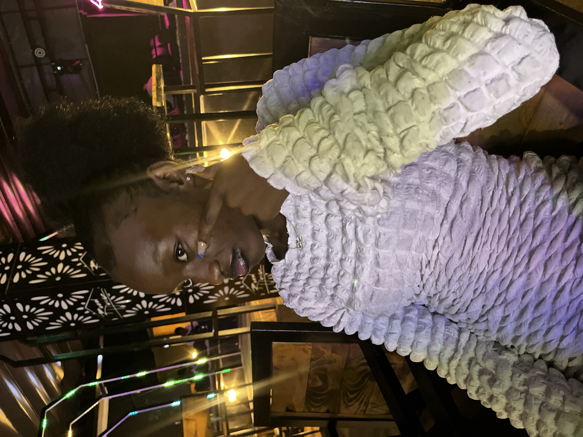 A young Black woman with dark skin and an afro hairstyle is seated in what appears to be an indoor restaurant or lounge with ambient lighting. She is looking to her left, with her right hand raised to her face, touching her cheekbone with her index finger. She is wearing a textured, multi-colored garment, transitioning from white to light yellow and purple. Her lips are dark, and she has a light blue accent on her cheekbone near her eye. In the background, there are wooden chairs, decorative metal screens with floral patterns, and colorful string lights. The overall scene suggests a social gathering or outing in Howayi, Nigeria.