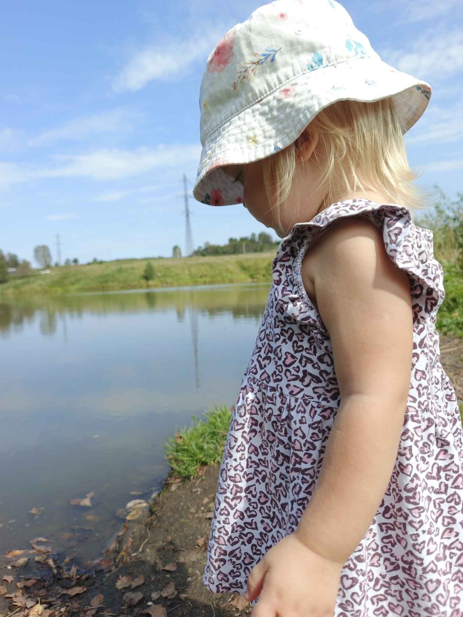 A young child with blonde hair, wearing a white hat with floral patterns and a patterned dress, is standing by the edge of a body of water. The child is looking down towards the water. The background shows a grassy slope, more trees, a power line tower, and a blue sky with some clouds. The location is Urtiushka, Russia.