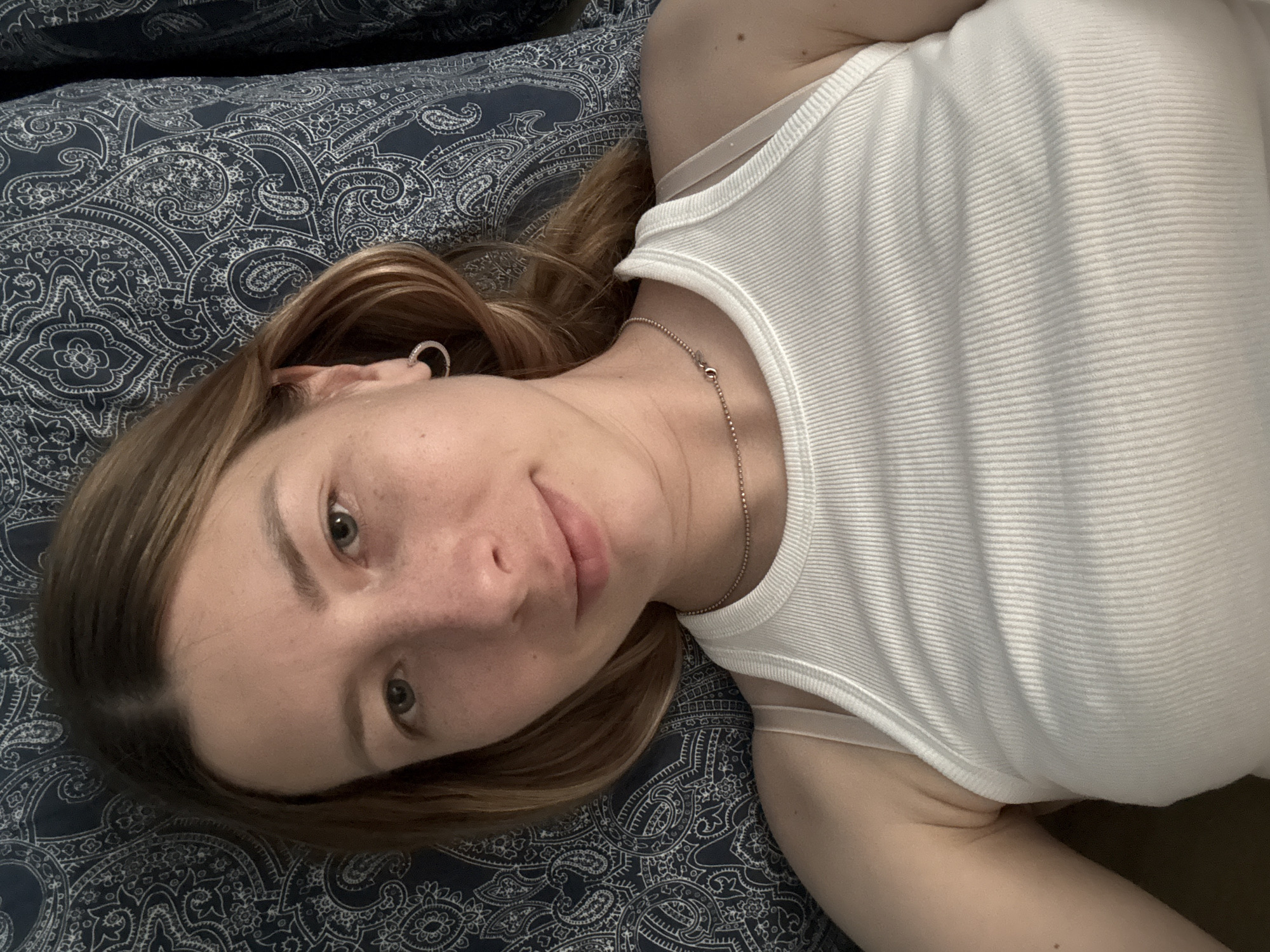 A fair-skinned person with light brown hair is lying on their back on a patterned blue and white surface, likely a bed or couch. They are wearing a white ribbed tank top and a silver chain necklace. An earring is visible in their left ear. The location of the image is not discernible as Moscow, Russia. There are no distinguishing landmarks or objects.