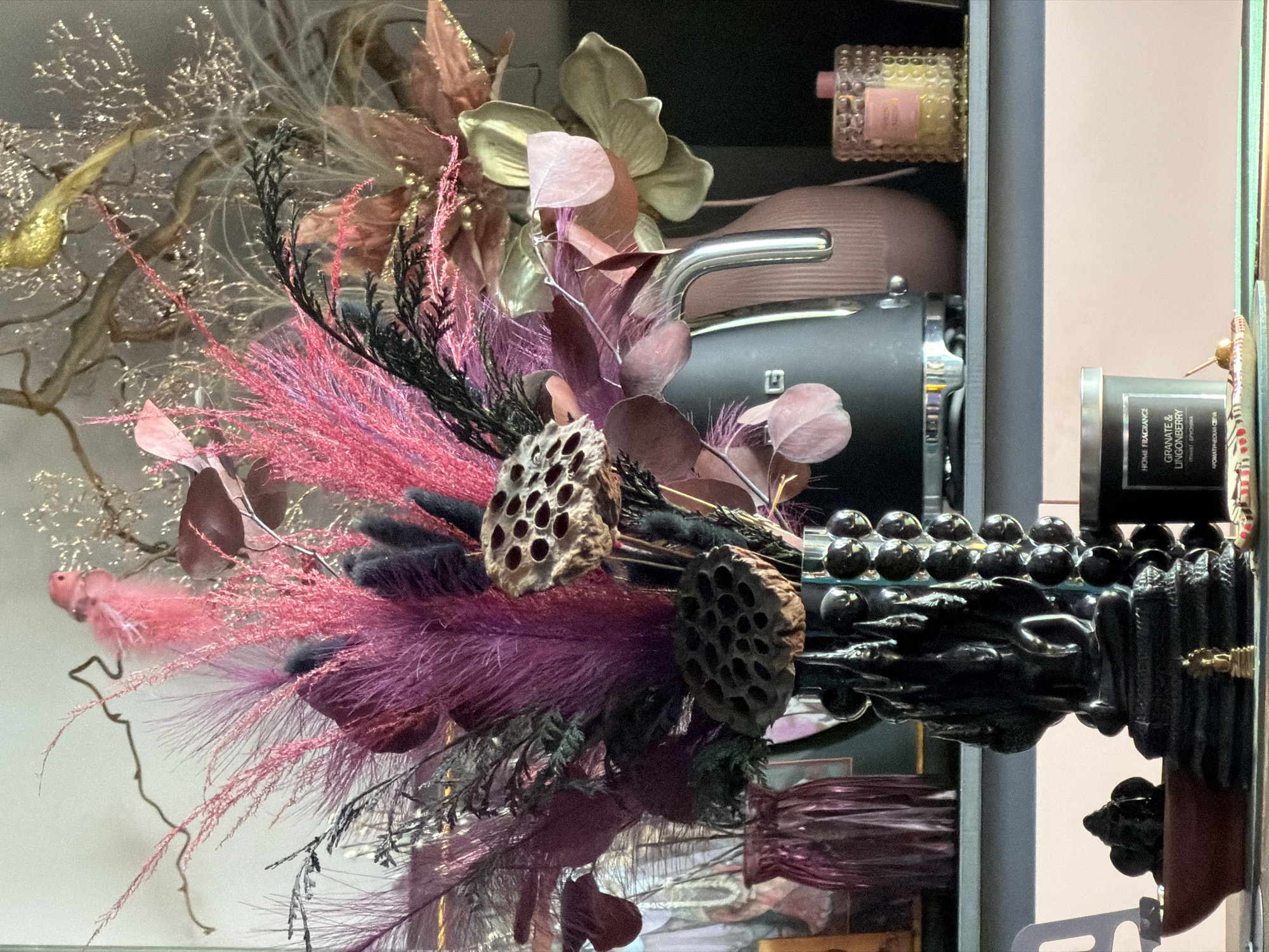 The image depicts a floral arrangement with pink and purple feathery plumes, dried lotus seed pods, and silver-colored branches. The arrangement is placed within a display cabinet or shelf unit. Other items are visible on the shelves, including a decorative perfume bottle, a pink container, and a candle in a dark gray jar labeled "HOME FRAGRANCE GRANATE & LINGONBERRY". The image appears to have been taken inside a store or decorative space. There is no evidence to suggest that this image comes from Moscow, Russia.