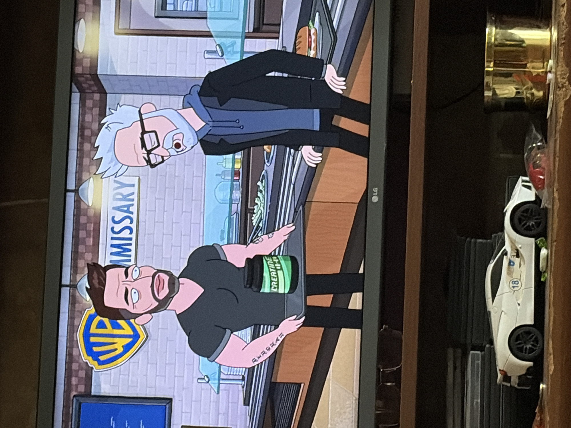 This image shows a screen displaying an animated scene with two cartoon characters in what appears to be a cafeteria. The character on the left is a man with dark hair and a beard, wearing a black t-shirt. He is holding a tray with a green container of "CREATINE" supplement. He has tattoos on his arms. The character on the right is an older man with light-colored spiky hair, wearing glasses, a blue hoodie, and a black blazer. He is standing near a food counter with a sandwich on a plate. Behind them, there is a Warner Bros. logo and a sign that reads "COMMISSARY." The bottom portion of the image shows a shelf with a white toy car, some DVDs, and a metallic gold container.