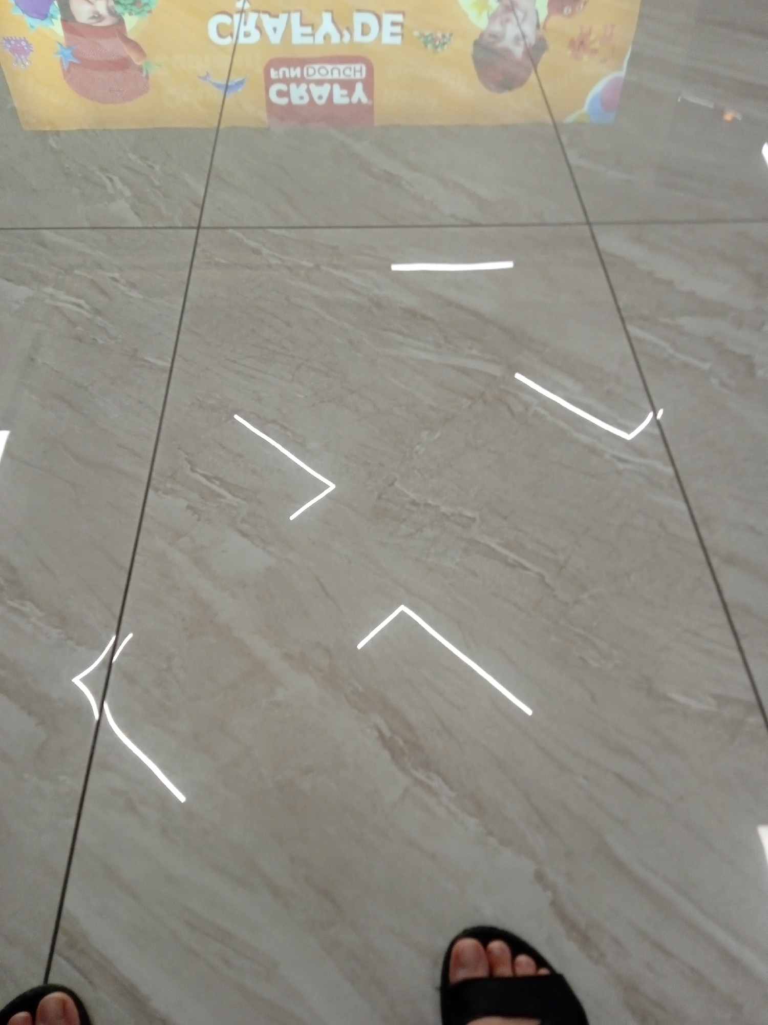 In Yenişehir, Turkey, the image captures a floor tiled with large, light-colored, marble-effect tiles. The lighting creates bright reflections on the glossy surface, forming white lines and shapes. A portion of a yellow banner is visible at the top, featuring text that appears to be "CEVLA, DE" and "CNU PONCH CEVLA" in a reversed, reflected manner. There are also reflections of cartoon-like figures on the banner, one with red hair and another with darker hair and a brown garment. Two feet are visible at the bottom of the frame; one wearing a black sandal is on the right, and another foot is partially visible on the left. The scene is viewed from a slightly elevated perspective looking down.