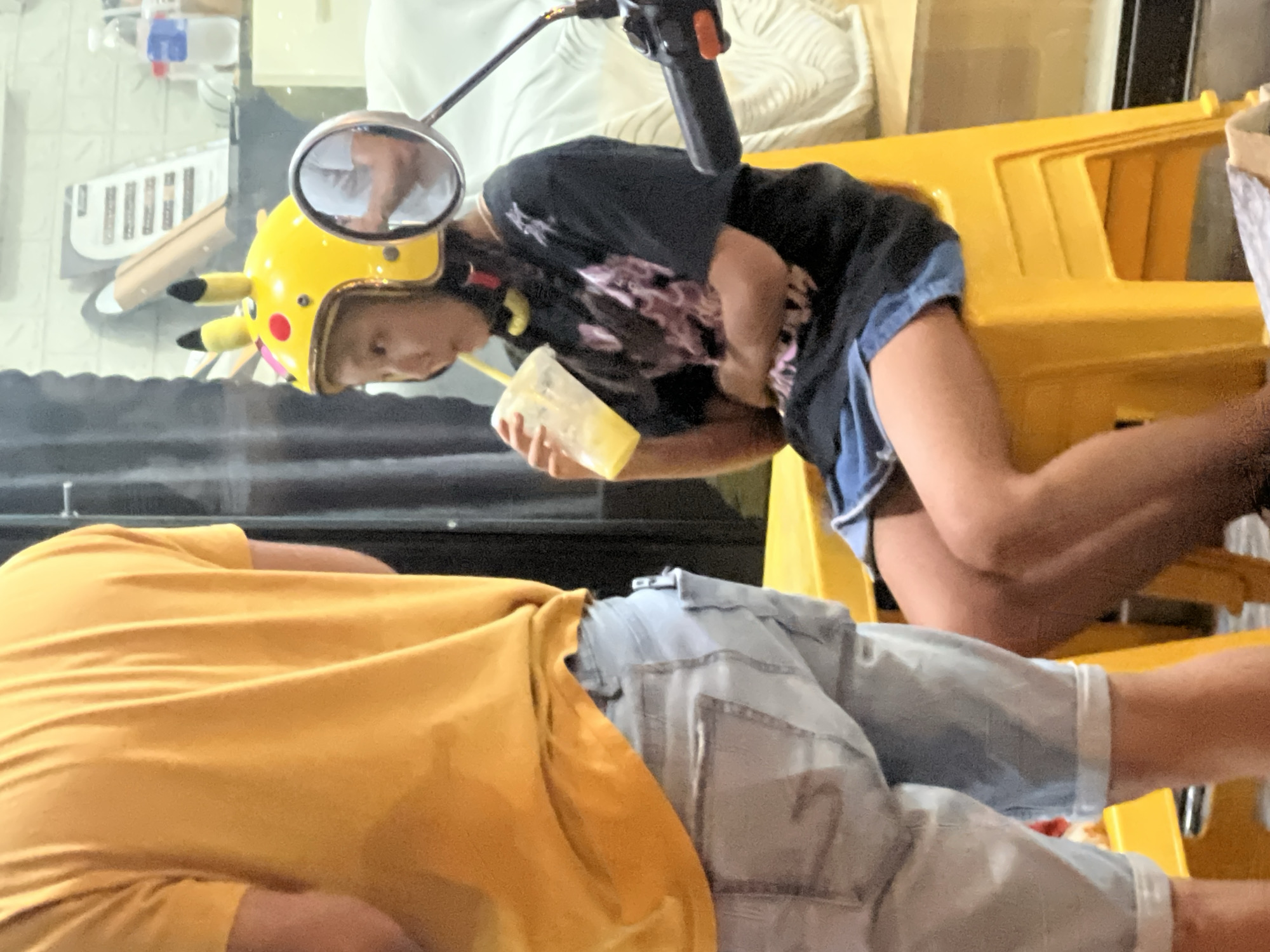 The image shows a young person wearing a yellow helmet with Pikachu ears, sitting on a yellow plastic chair. They are holding a cup with a yellow straw and appear to be drinking. The person is wearing a black t-shirt with a graphic print and blue denim shorts. To the left, another person is visible from the waist down, wearing a yellow t-shirt and light blue denim shorts. The background suggests an indoor environment, possibly a cafe or shop, with a reflective surface that shows a partial view of another person. There are shelves and bottles in the background. The overall lighting is dim, with some artificial light sources. The scene is likely set in the city of Đà Nẵng, Vietnam, given the context.