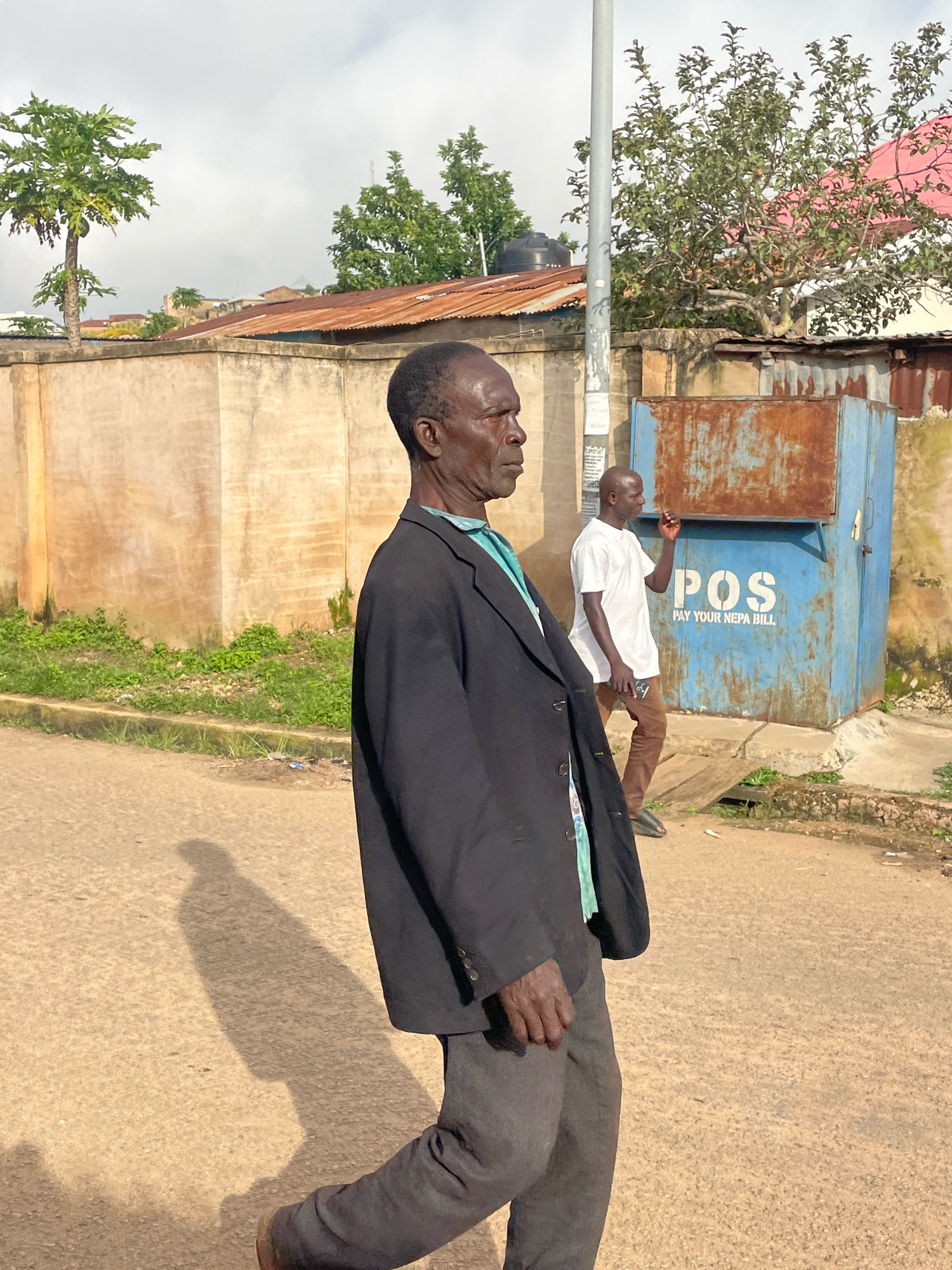 In Jos, Nigeria, on a street with a sandy-colored asphalt surface, two men are walking. The man in the foreground, viewed from the side in profile, is wearing a black suit jacket over a light blue collared shirt and dark grey trousers. His left hand is held near his waist. Behind him and to the right, a second man, bald and wearing a white t-shirt and brown trousers, is also walking. He is holding something to his mouth, possibly a cigarette. They are walking past a large, light blue metal structure with "POS PAY YOUR NEPA BILL." written on it in white letters. To the left of the POS structure, there is a rusty metal object. A grey utility pole stands behind the two men. In the background, there is a concrete wall with an orange corrugated metal roof on top. Beyond the wall, trees and parts of buildings are visible under a cloudy sky. Green grass and weeds grow along the side of the road. The shadow of the man in the foreground is cast on the road to his left.