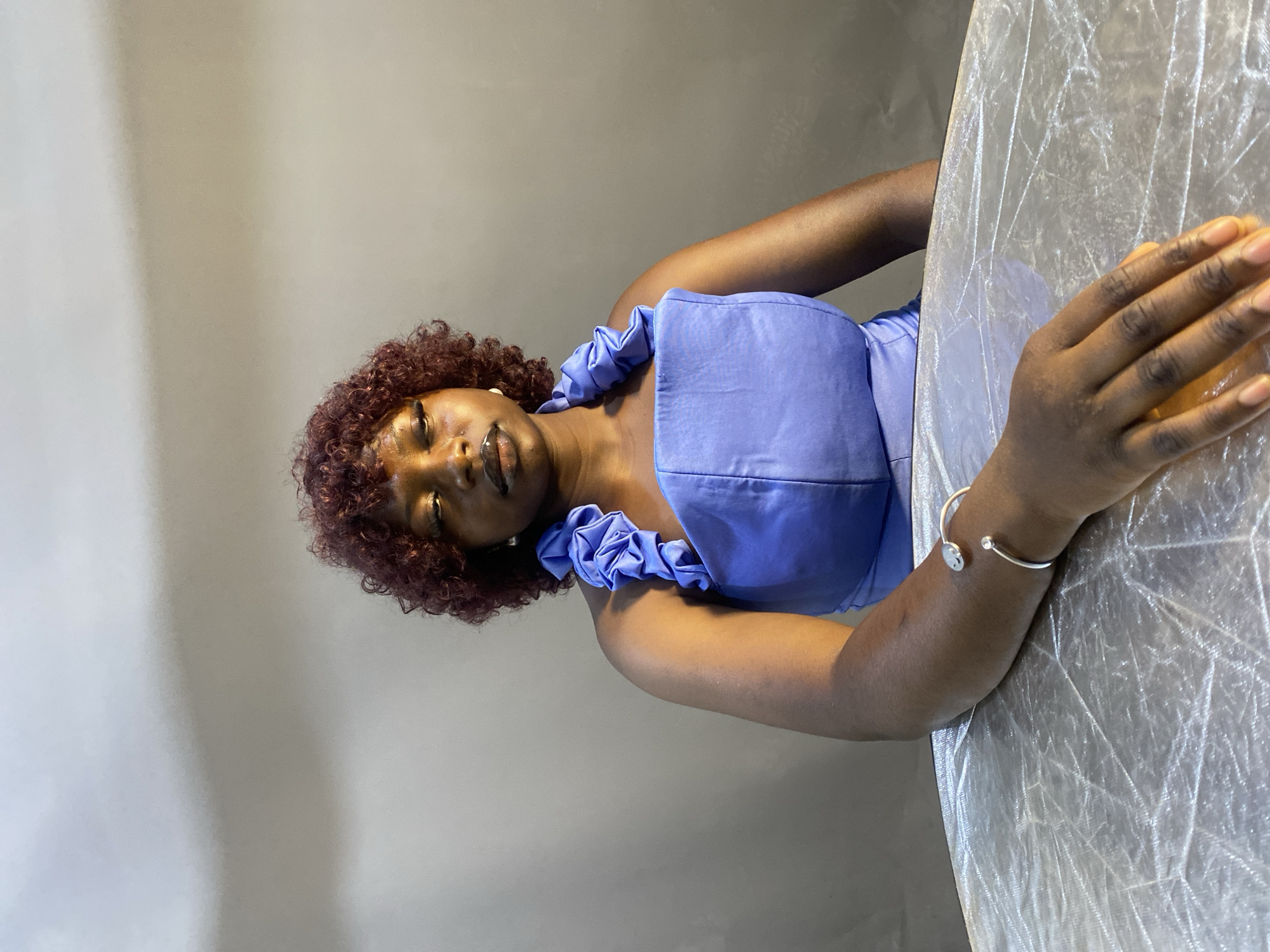A medium shot shows a woman with reddish-brown curly hair and dark skin sitting at a table covered with a crinkled, reflective material. She is wearing a blue strapless top with ruffled shoulder straps. Her right arm rests on the table, and she is wearing a silver bracelet on her wrist. She has makeup on, including dark lipstick and eyeliner. The background is a plain gray wall. The lighting creates shadows on her face and the wall. The overall scene appears to be indoors, possibly in a studio, and there is no indication of a specific location in Jos, Nigeria.