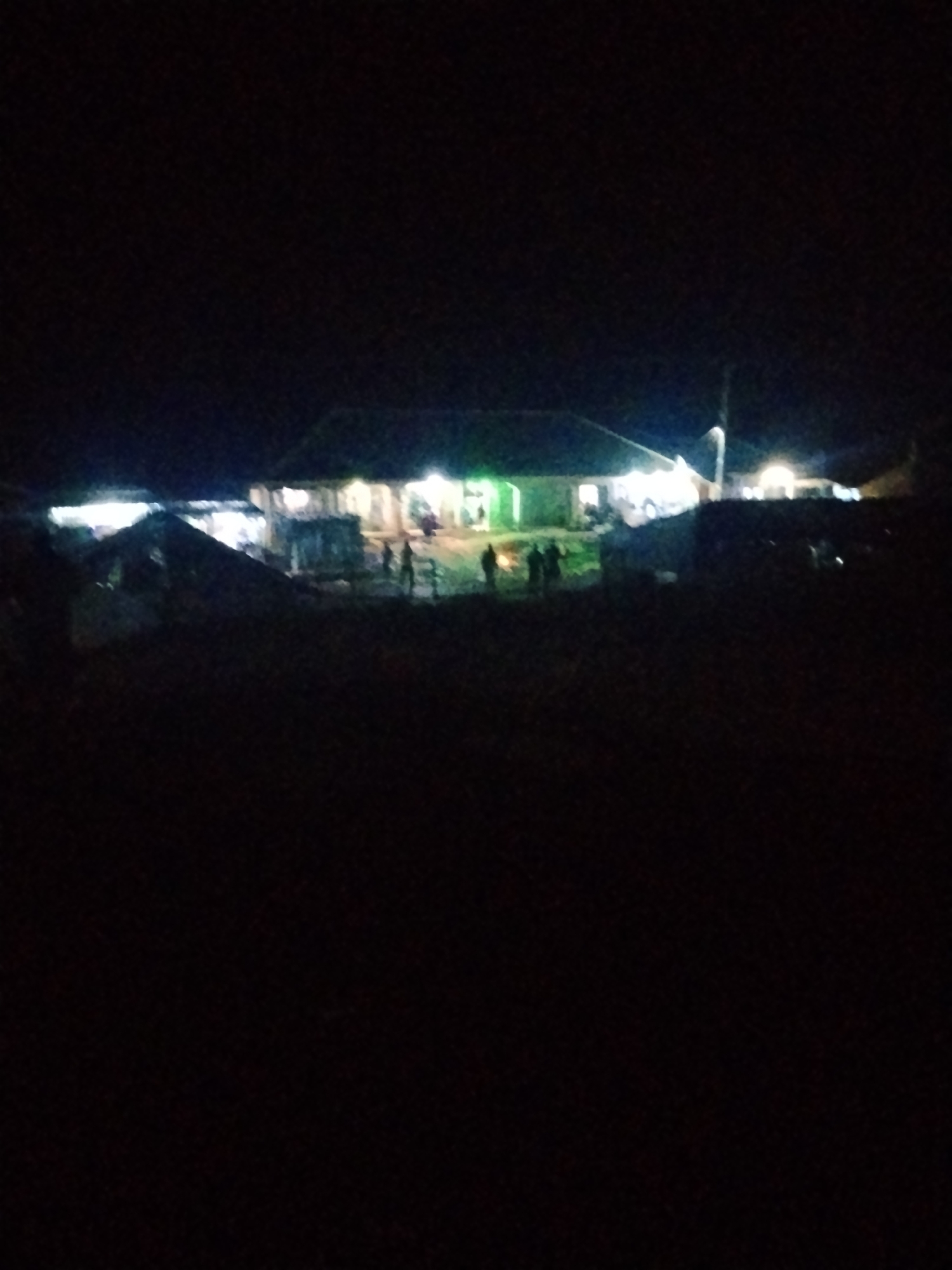 The image depicts a dimly lit outdoor scene at night. In the background, there is a building with illuminated windows, suggesting it is occupied or operational. Several figures, appearing as silhouettes, are visible in front of and around the building. The foreground is predominantly dark, with some blurry shapes that could be vegetation or uneven terrain. The overall impression is of a gathering or activity taking place outdoors after dark, possibly in a semi-rural or less developed area of Durumi, Nigeria, given the low light conditions and the appearance of the structures.