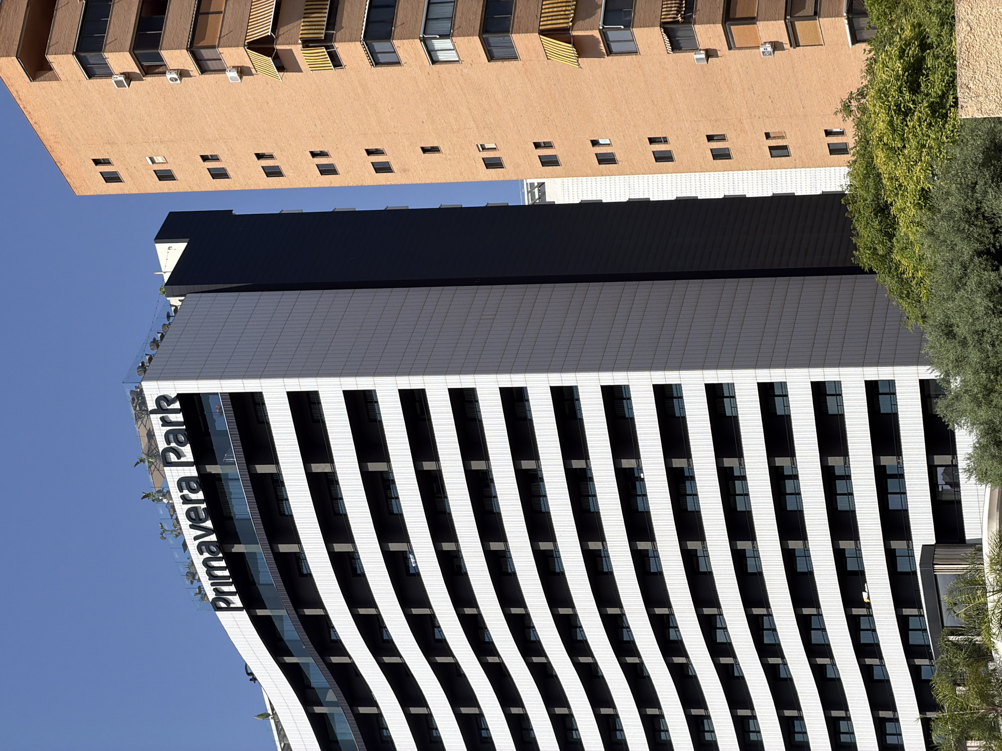 The image shows two tall buildings in Benidorm, Spain. The building on the left is modern and white with horizontal dark gray balconies, featuring the name "Primavera Park" in large black letters at the top. It has a curved facade. To its right, a taller, older building constructed of orange brick is visible. This building has numerous windows and several air conditioning units mounted on its exterior. Yellow awnings are visible on some of the balconies of the brick building. Green trees and foliage are in the foreground, obscuring the lower portions of the buildings. The sky is clear and blue.