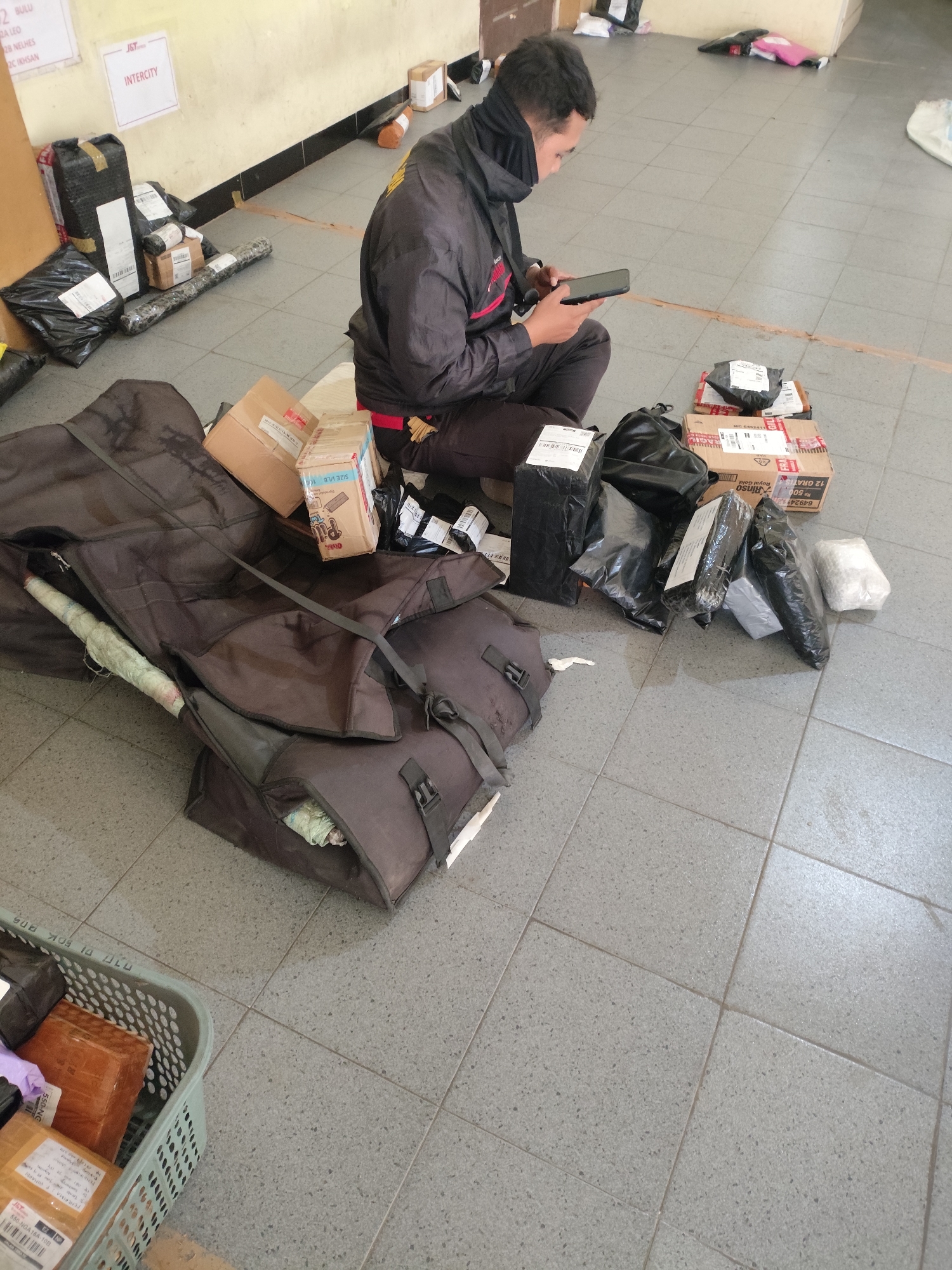 In Petok, Indonesia, a man in a black jacket and dark pants sits on the floor amidst a collection of packages and large dark bags. He is looking at a smartphone in his hands. The packages are of various sizes and shapes, some are wrapped in black plastic, others are in cardboard boxes. One large rolled item, wrapped in light-colored plastic, is partially visible under a dark bag. In the background, there are two white signs on the wall. One sign has "INTERCITY" printed on it in blue letters. The floor is tiled in a gray, speckled pattern. To the left of the frame, a light blue plastic basket contains several more packages. The scene appears to be an indoor waiting area or sorting facility for deliveries.